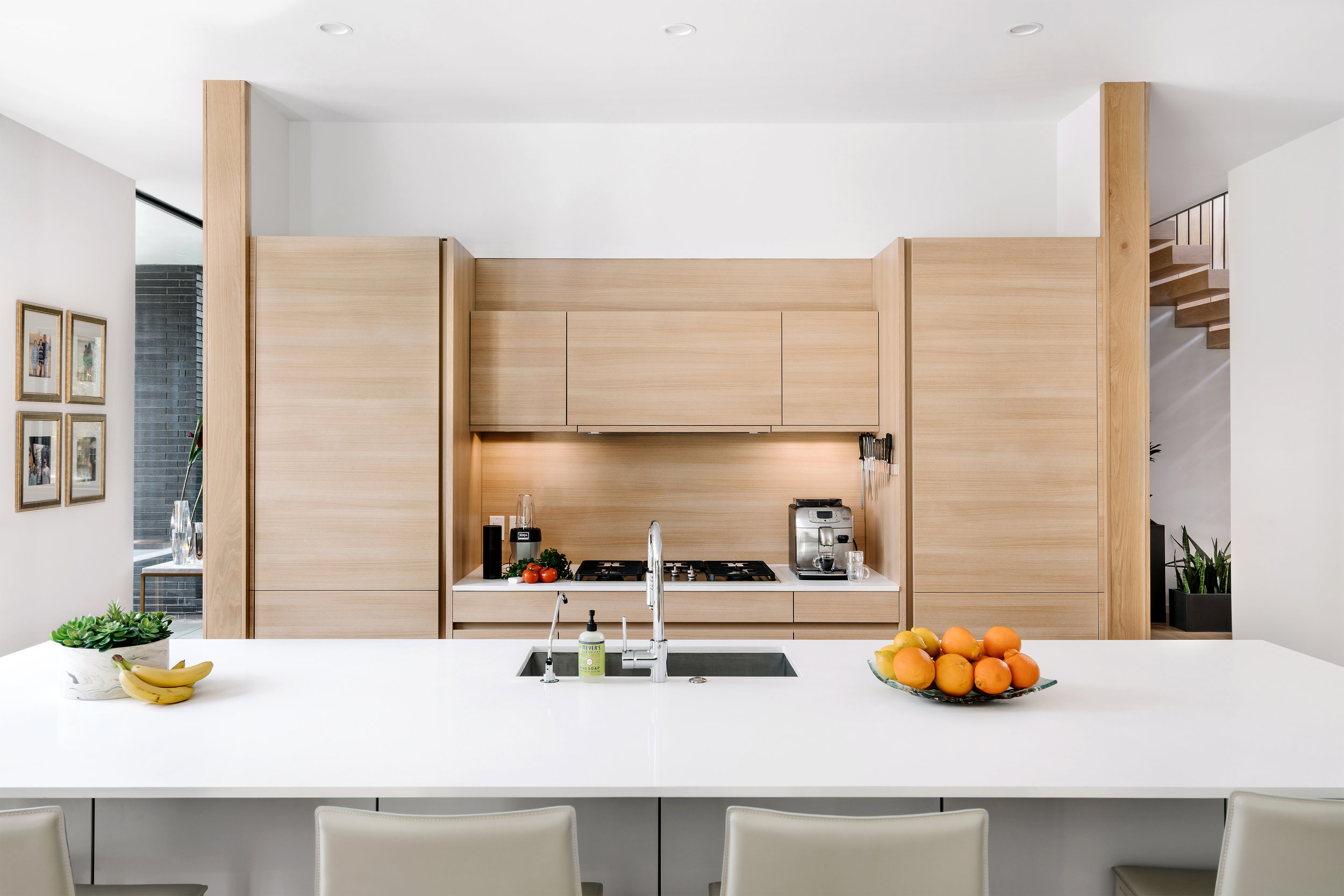 Modern kitchen in the Pecos Residence with wooden cabinets, white island countertop, fruits, coffee maker, and framed pictures on the wall.