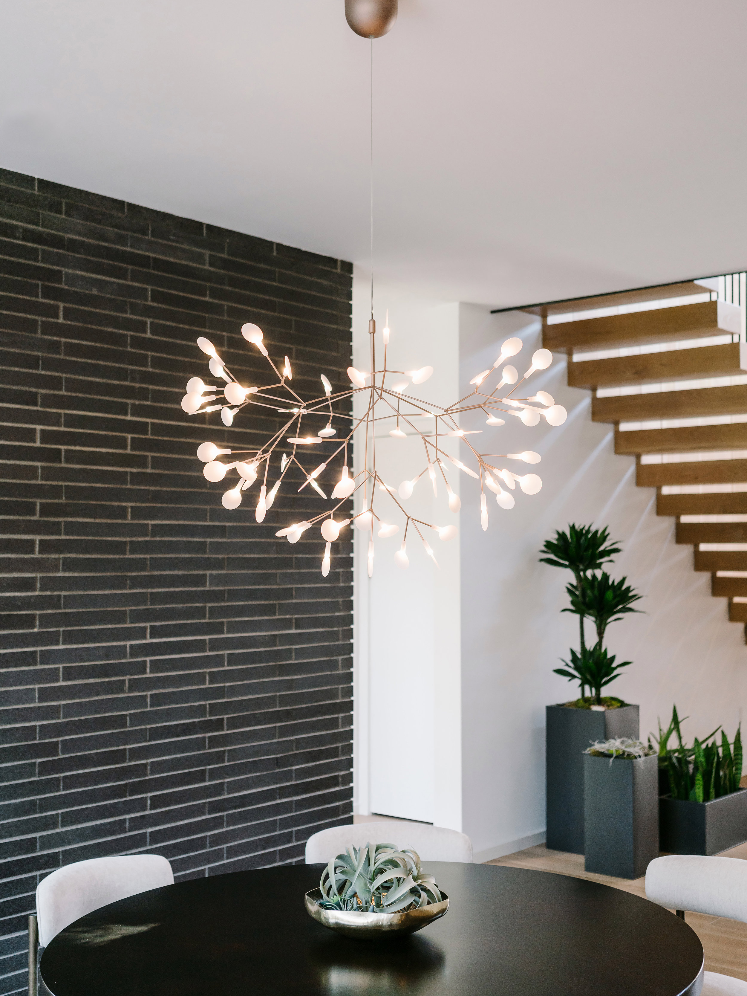 Modern dining area in the Pecos Residence with a black table, contemporary chandelier, and staircase. Potted plants adorn the space.