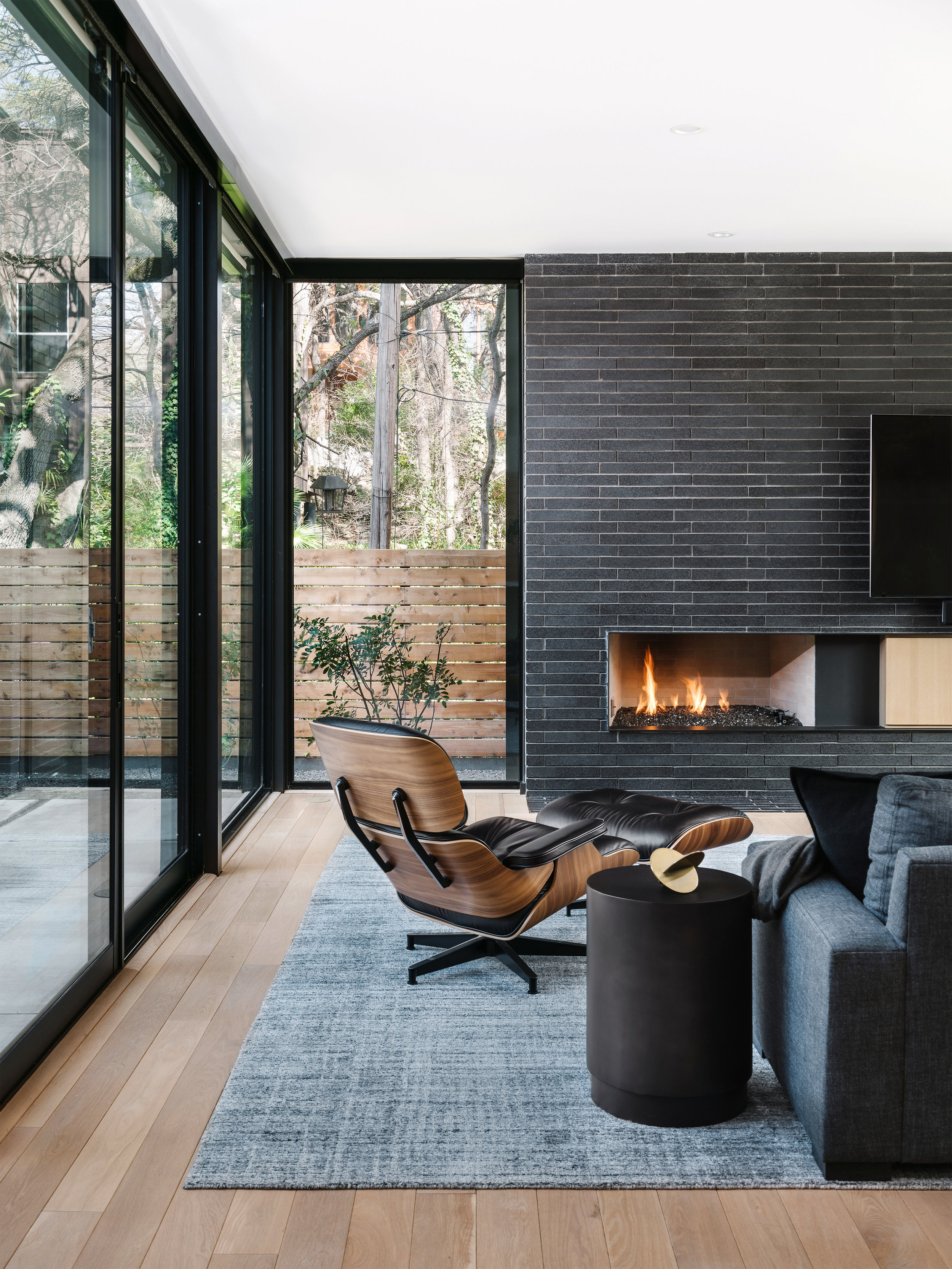 Modern living room in the Pecos Residence with a sleek fireplace, wooden armchair, cozy sofa, and large windows overlooking a wooded area.
