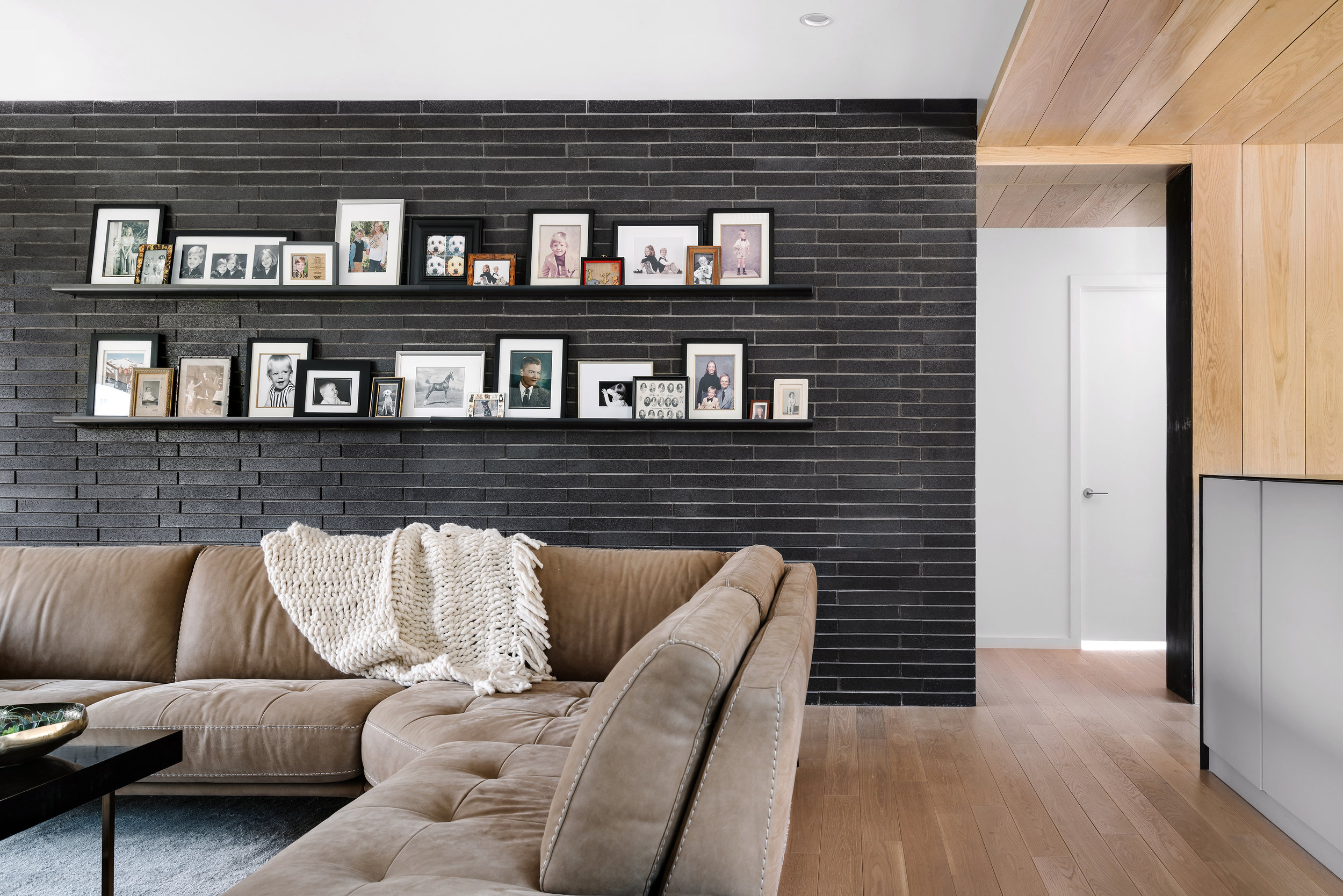A modern living room in the Pecos Residence with a brown sectional sofa, black brick wall displaying framed photos on shelves, and a cozy throw blanket.