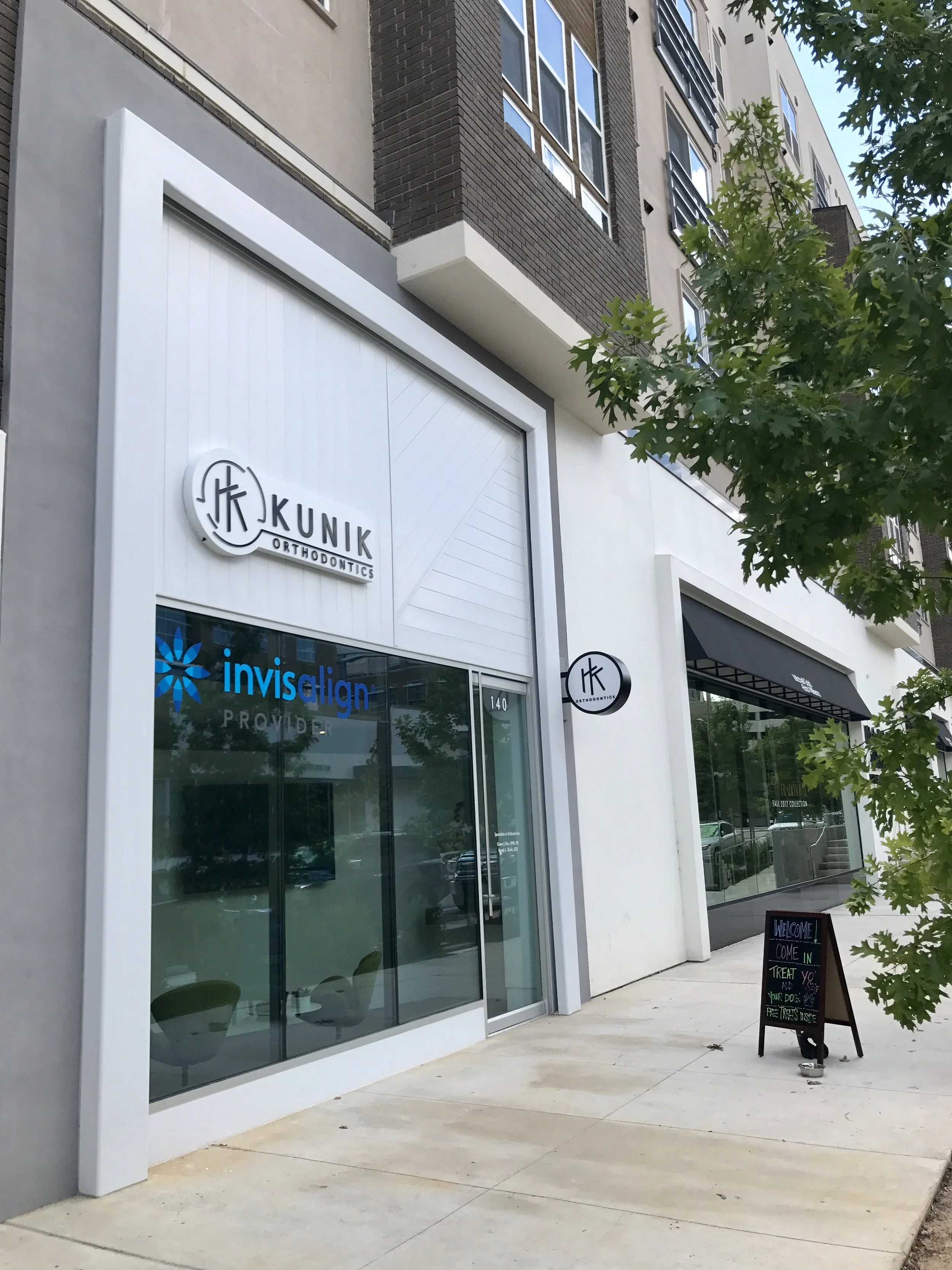  Exterior of Kunik Orthodontics with an Invisalign sign, light gray facade, sidewalk, and a chalkboard sign near the entrance. 