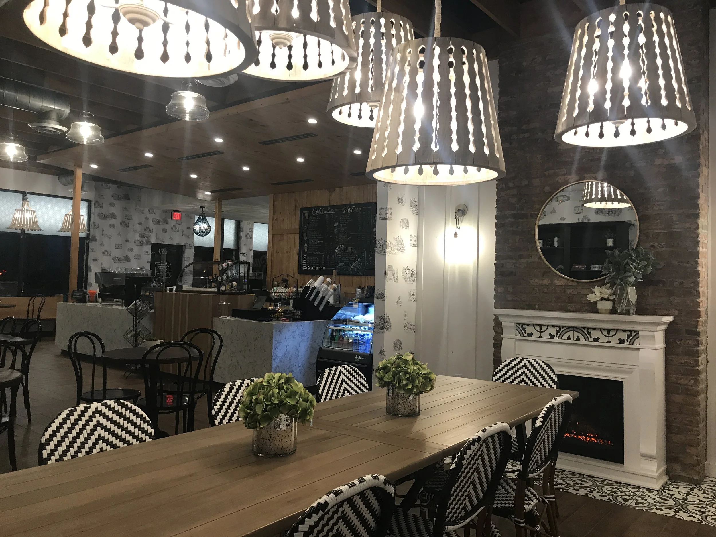  Interior of Summer Moon Coffee Bee Cave with wooden tables, modern pendant lights, black-and-white chairs, a fireplace, and a chalkboard menu. 
