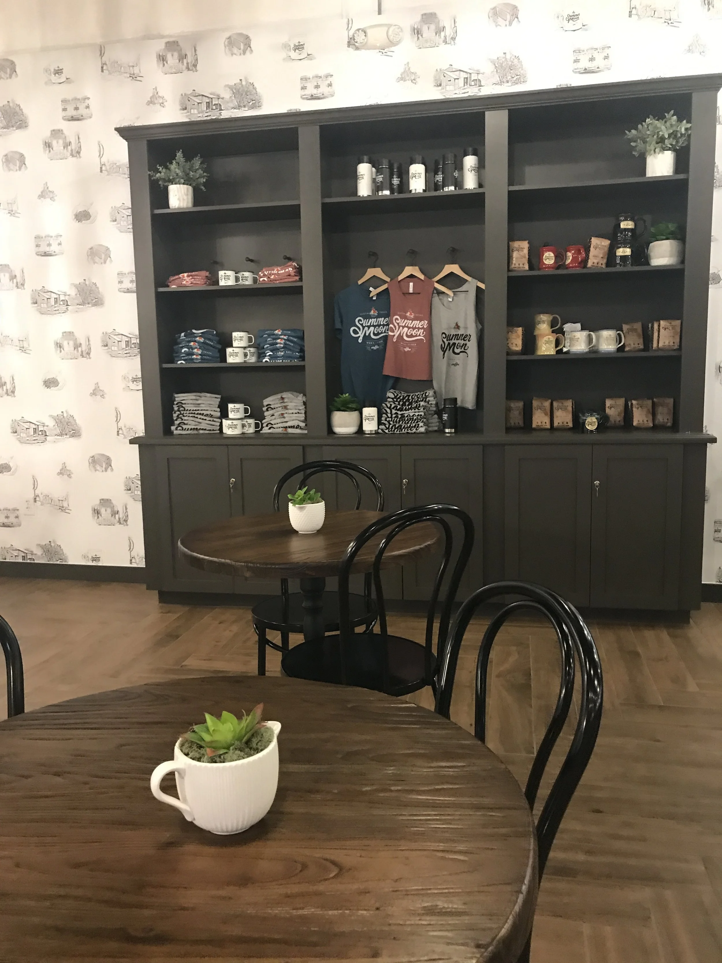  Summer Moon Coffee Bee Cave interior with dark wood and black furniture, potted plants on tables, and shelves displaying mugs, shirts, and other merchandise. 