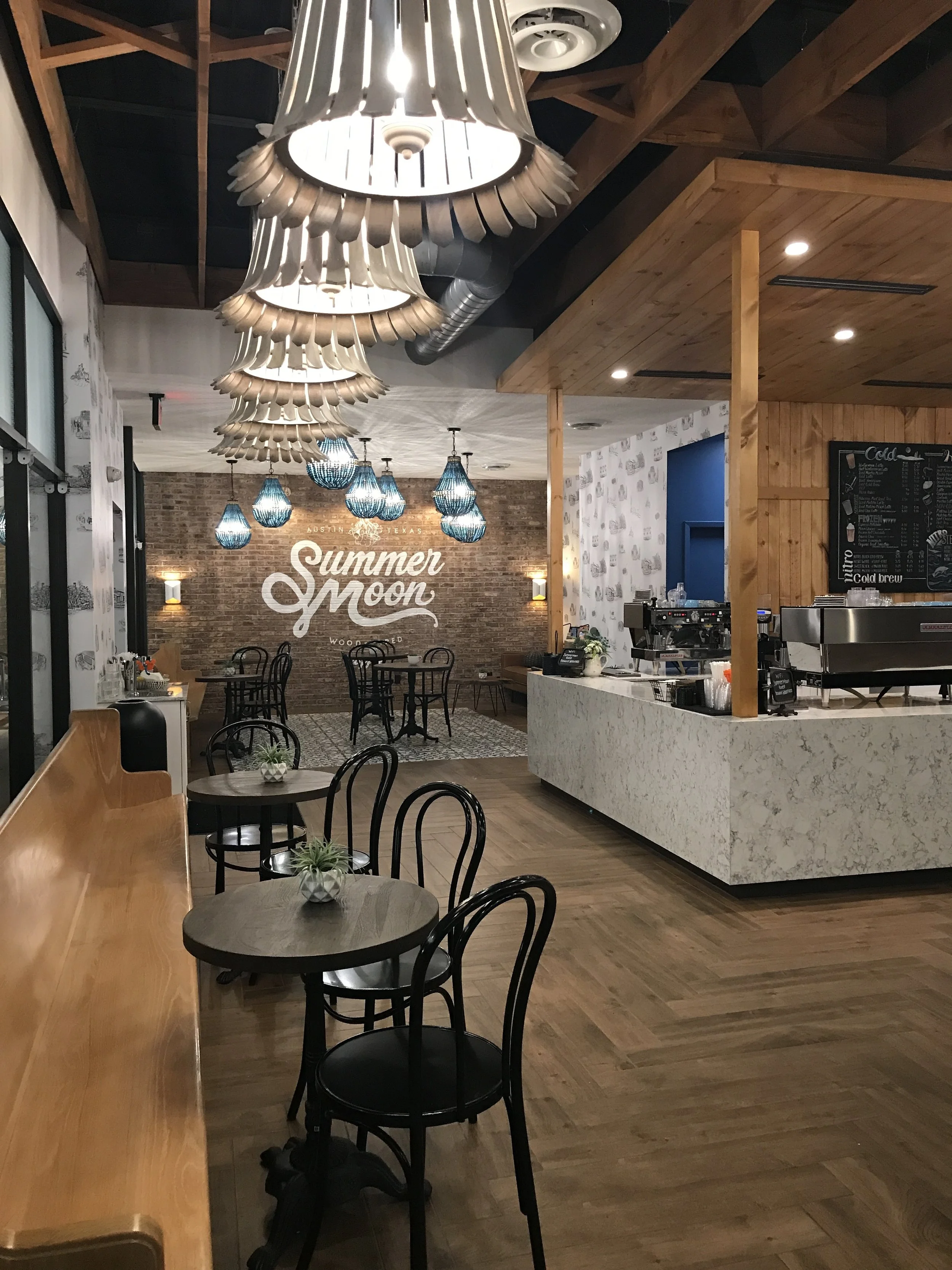  Interior of Summer Moon Coffee Bee Cave with wooden decor, round tables, a counter, and "Summer Moon" sign on a brick wall. 