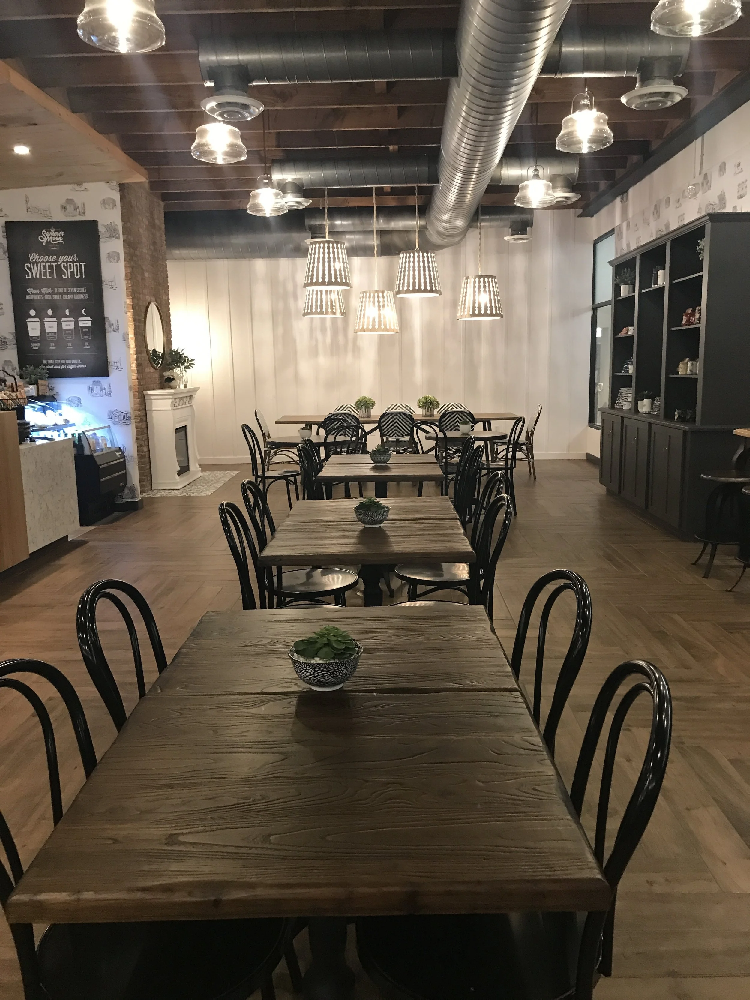  Summer Moon Coffee Bee Cave interior with wooden tables, black chairs, hanging lights, and a merchandise display cabinet in the background. 