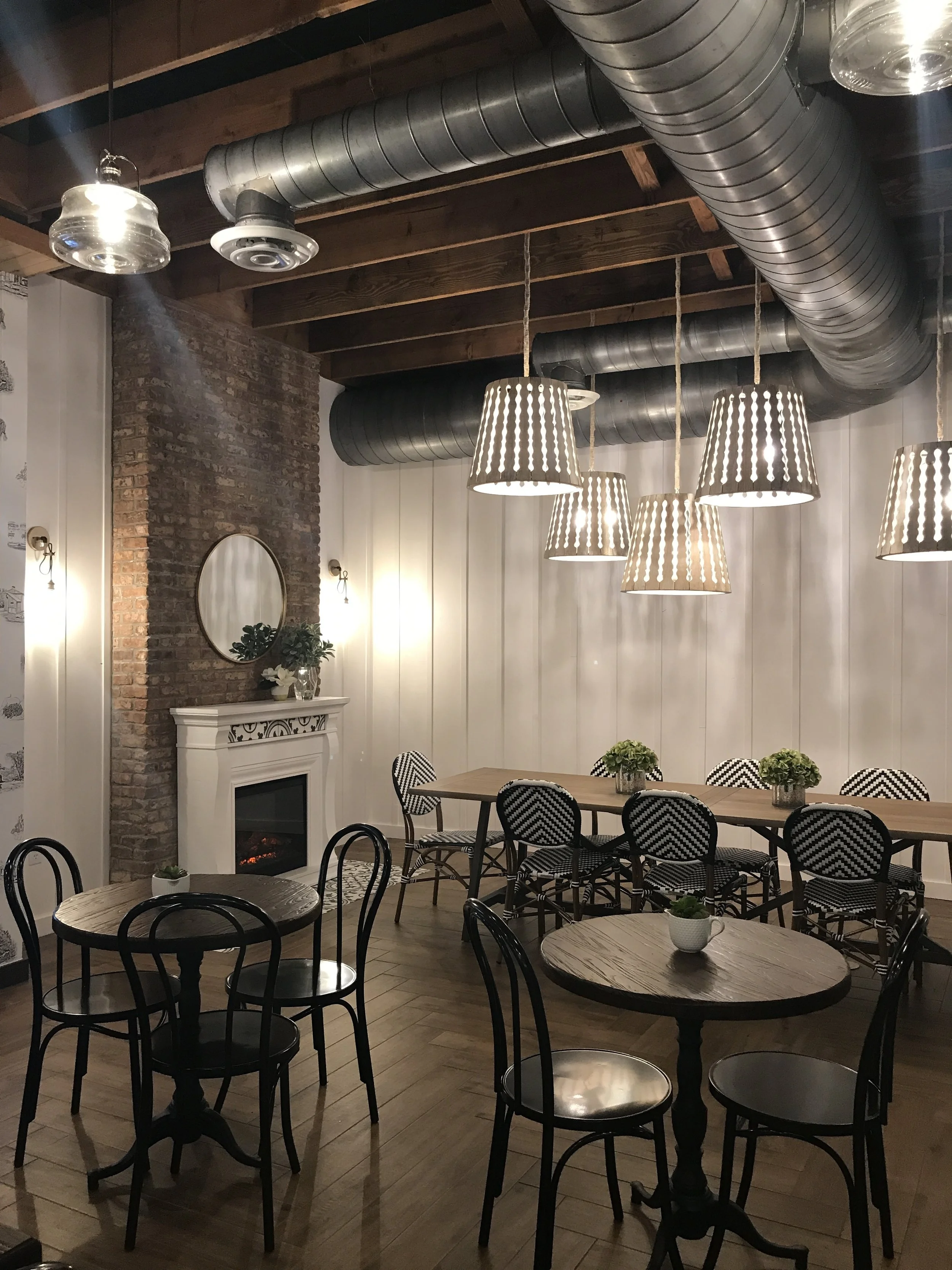  Summer Moon Coffee Bee Cave interior with hanging light fixtures, a fireplace, round tables, black chairs, and greenery on tables under large ducts. 