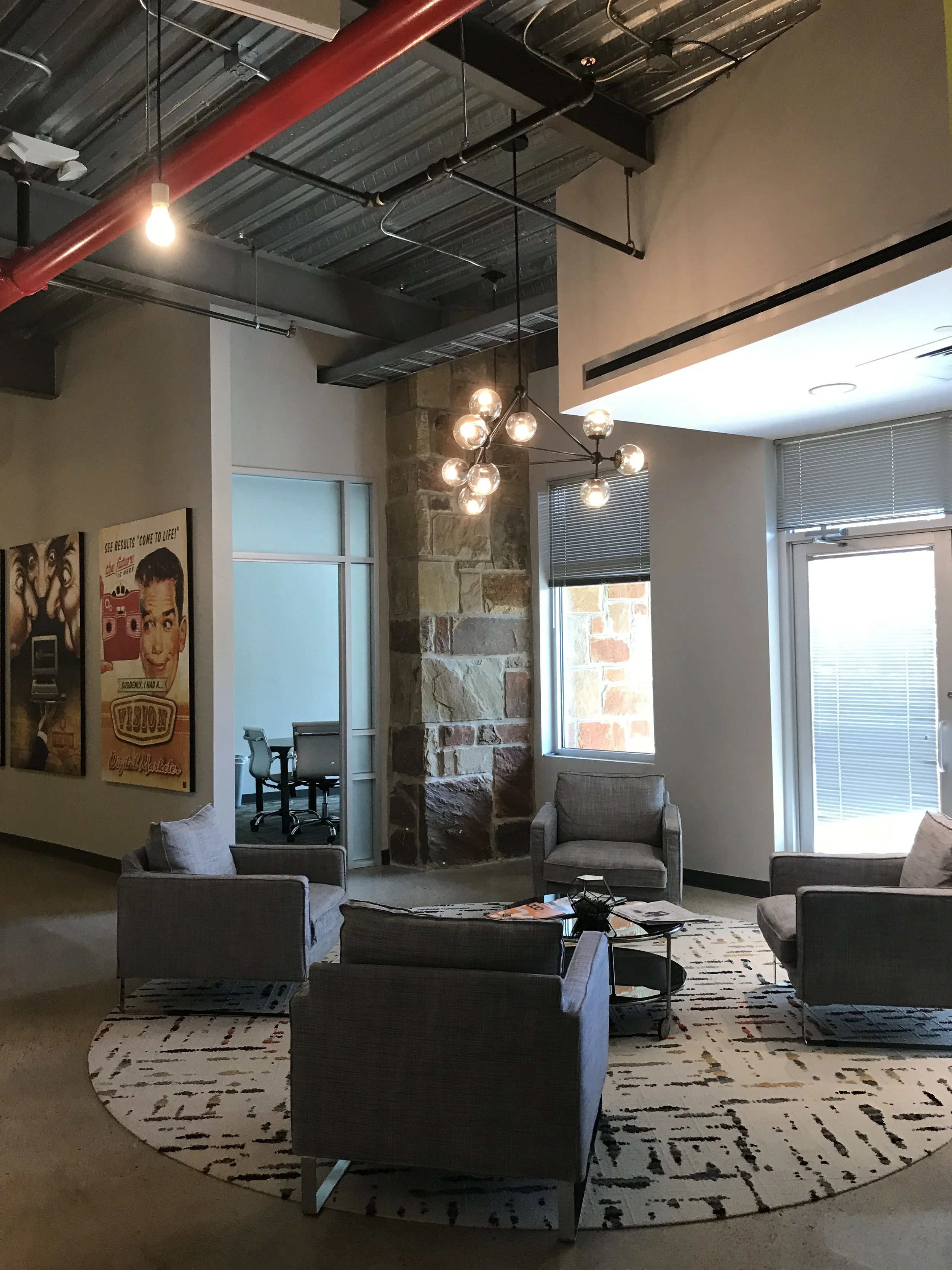  A Digital Marketer office lounge with gray chairs, a round rug, and a contemporary chandelier, adjacent to a small meeting room. 