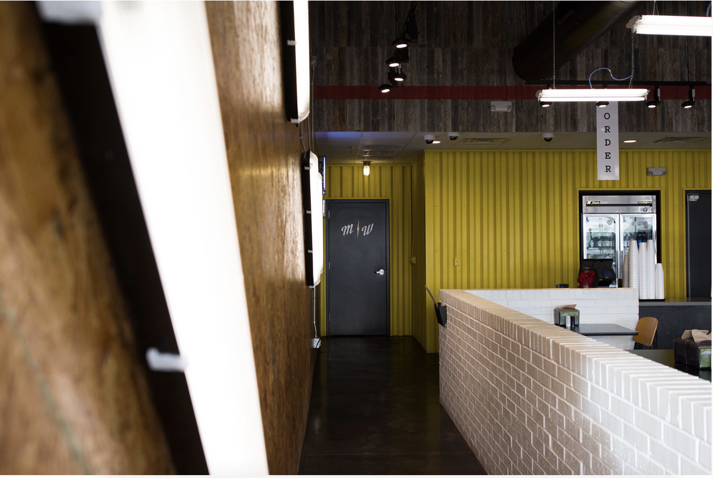 Interior of Flash Burger restaurant with a yellow wall, light brick partition, and an "Order" sign above a counter with a refrigerator behind.