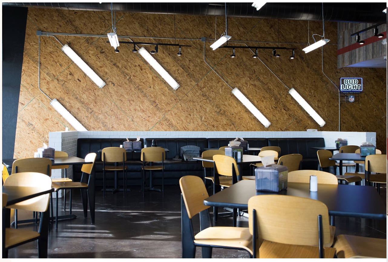 Flash Burger restaurant dining area with wooden chairs, tables, and multiple fluorescent tube lights, some hanging and others attached to a wooden accent wall.