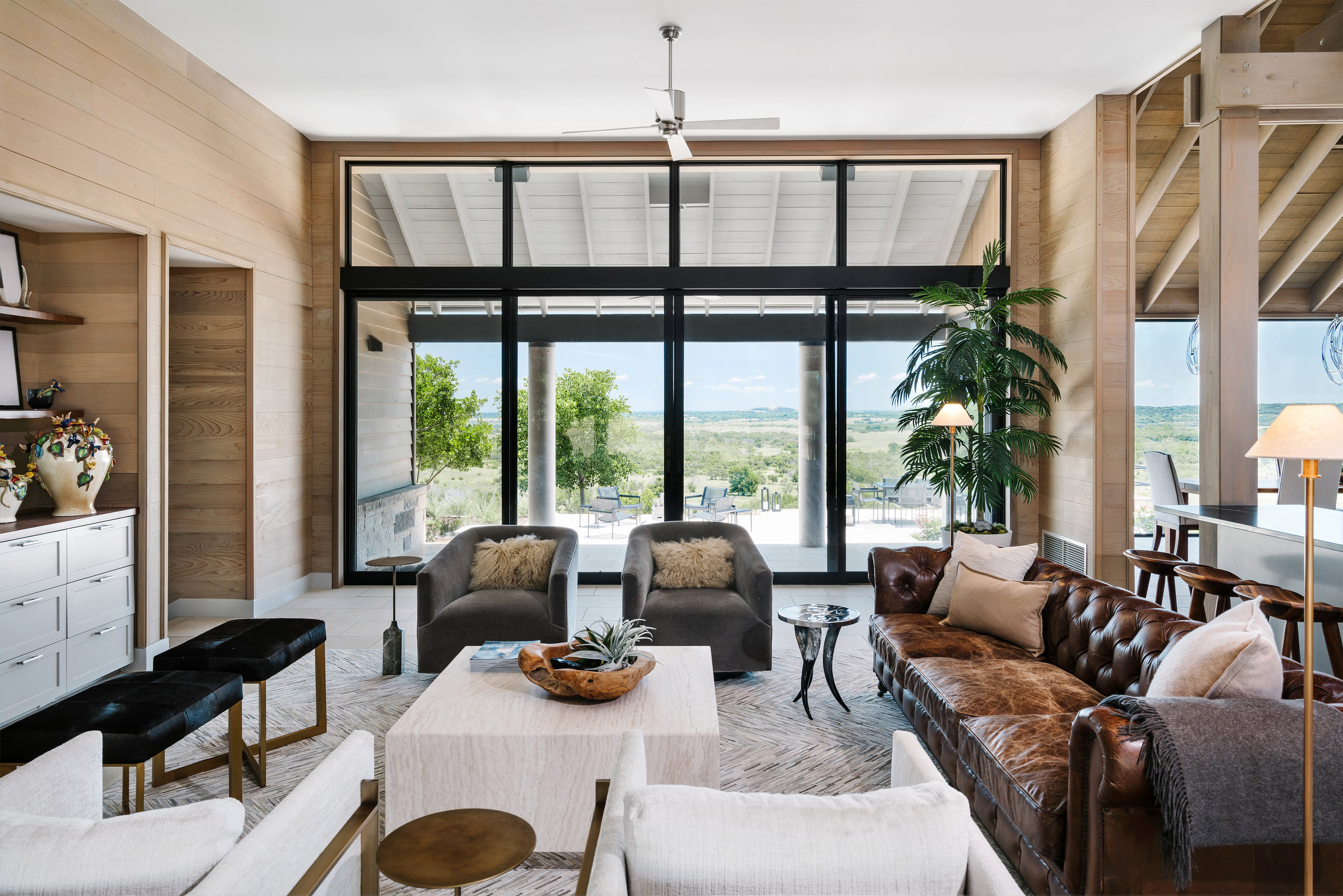 Spacious living room in a Lampassas ranch home with modern furniture, large windows, lush greenery outside, and a mix of earthy and neutral tones.