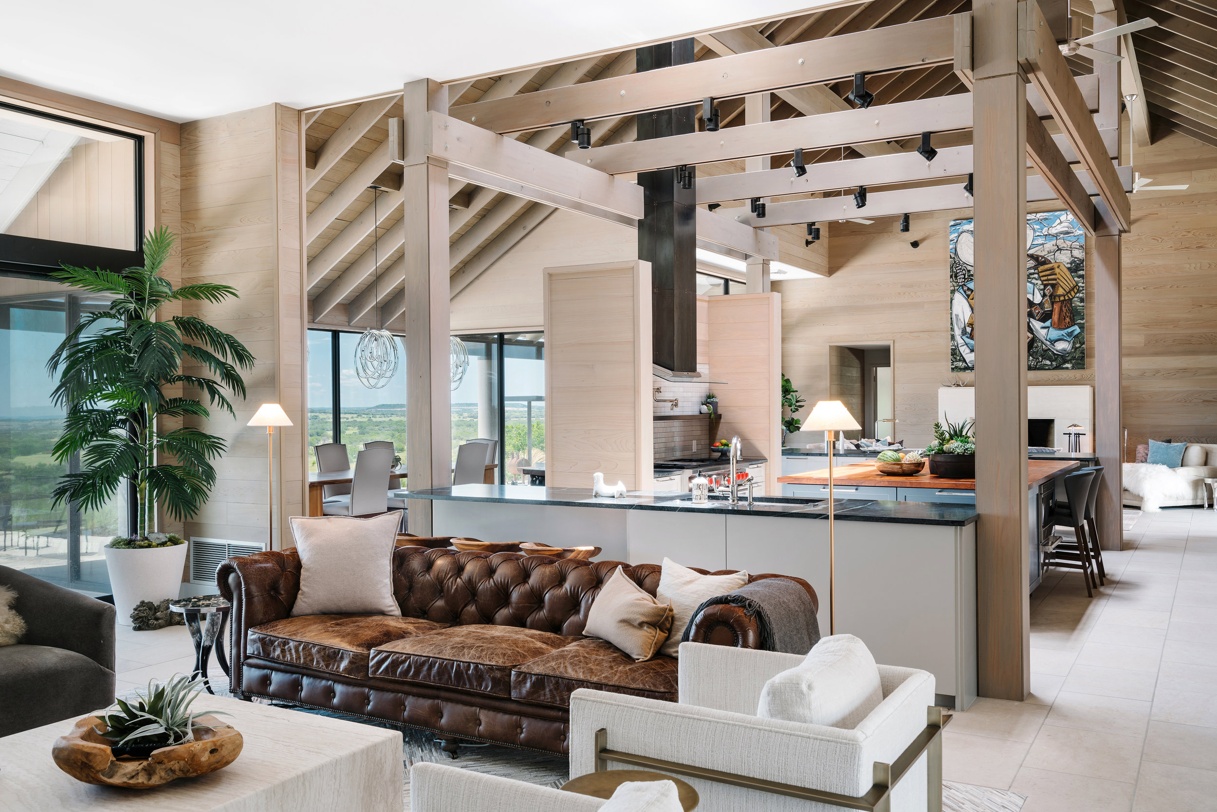 Open-concept living area in a Lampassas ranch home with a brown leather sofa, kitchen island, dining table, and large windows with scenic views.