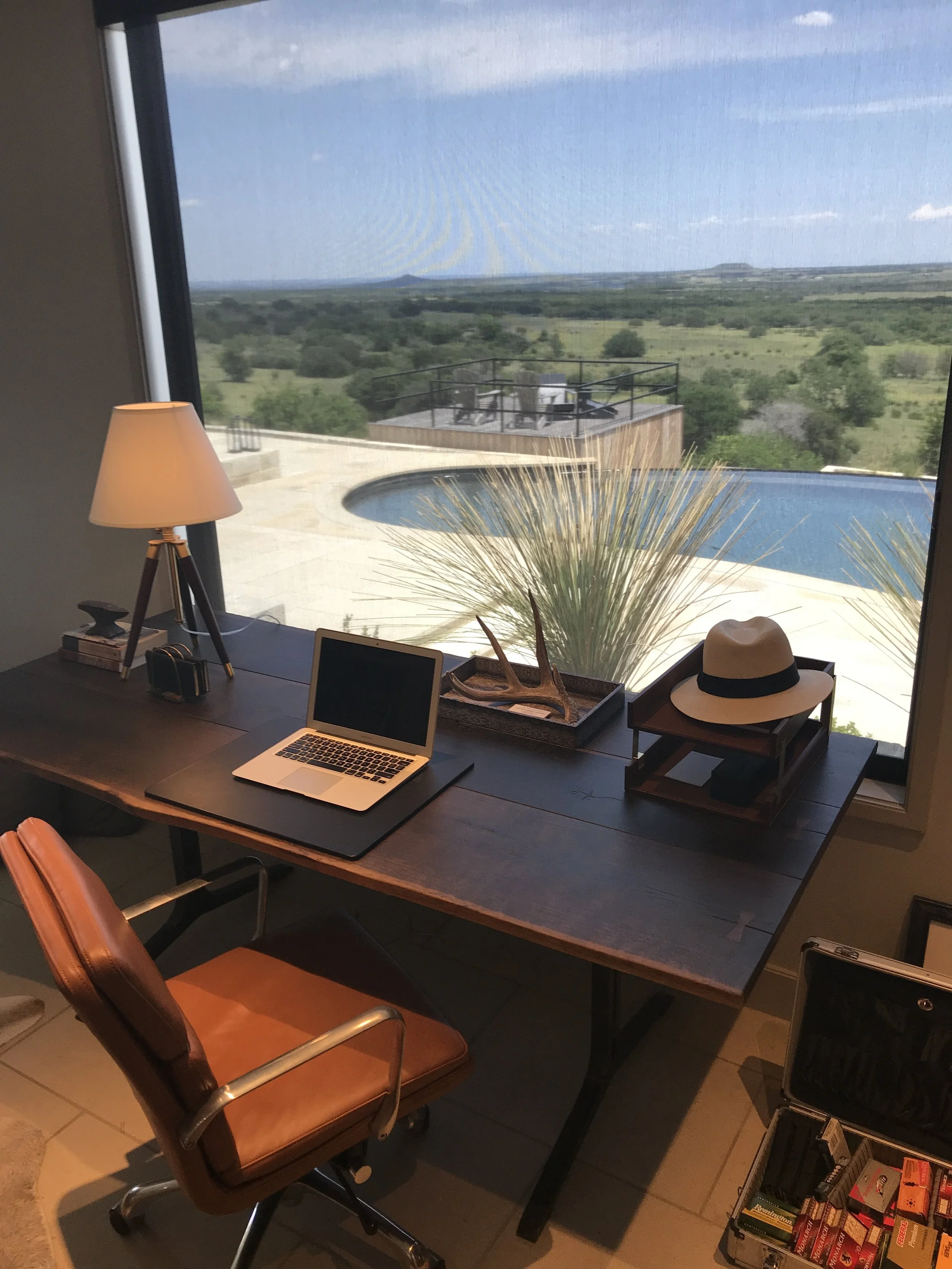  Office in a Lampassas ranch home with a desk, laptop, lamp, and hat, facing a large window overlooking a pool and scenic outdoor landscape. 