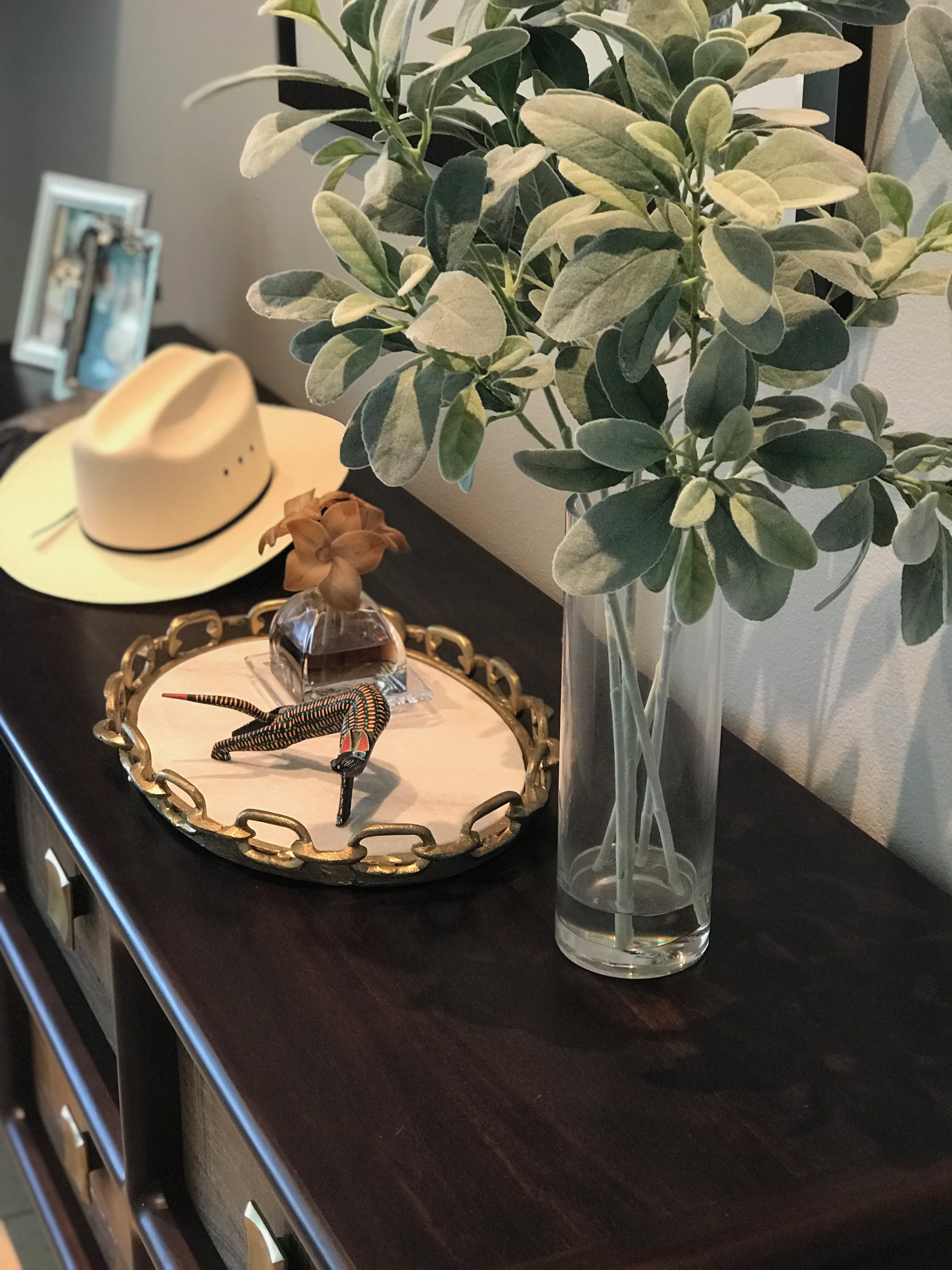Dark wooden dresser with a vase of green foliage, a decorative tray with items, and a cowboy hat in the background.