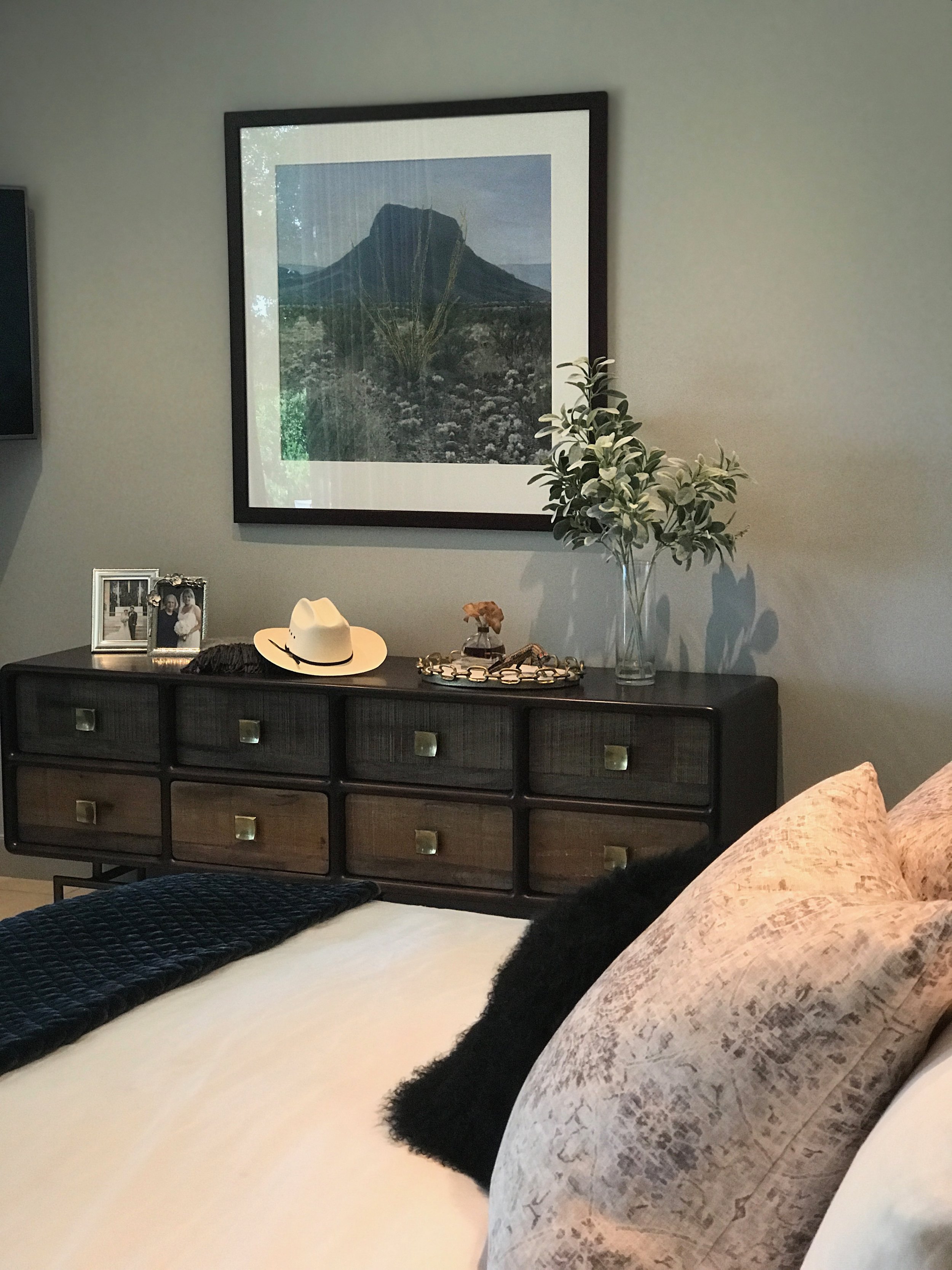 A bedroom in a Lampassas ranch home featuring a dresser with decor items, a framed photograph, a hat, and a vase, with the edge of a bed in view.