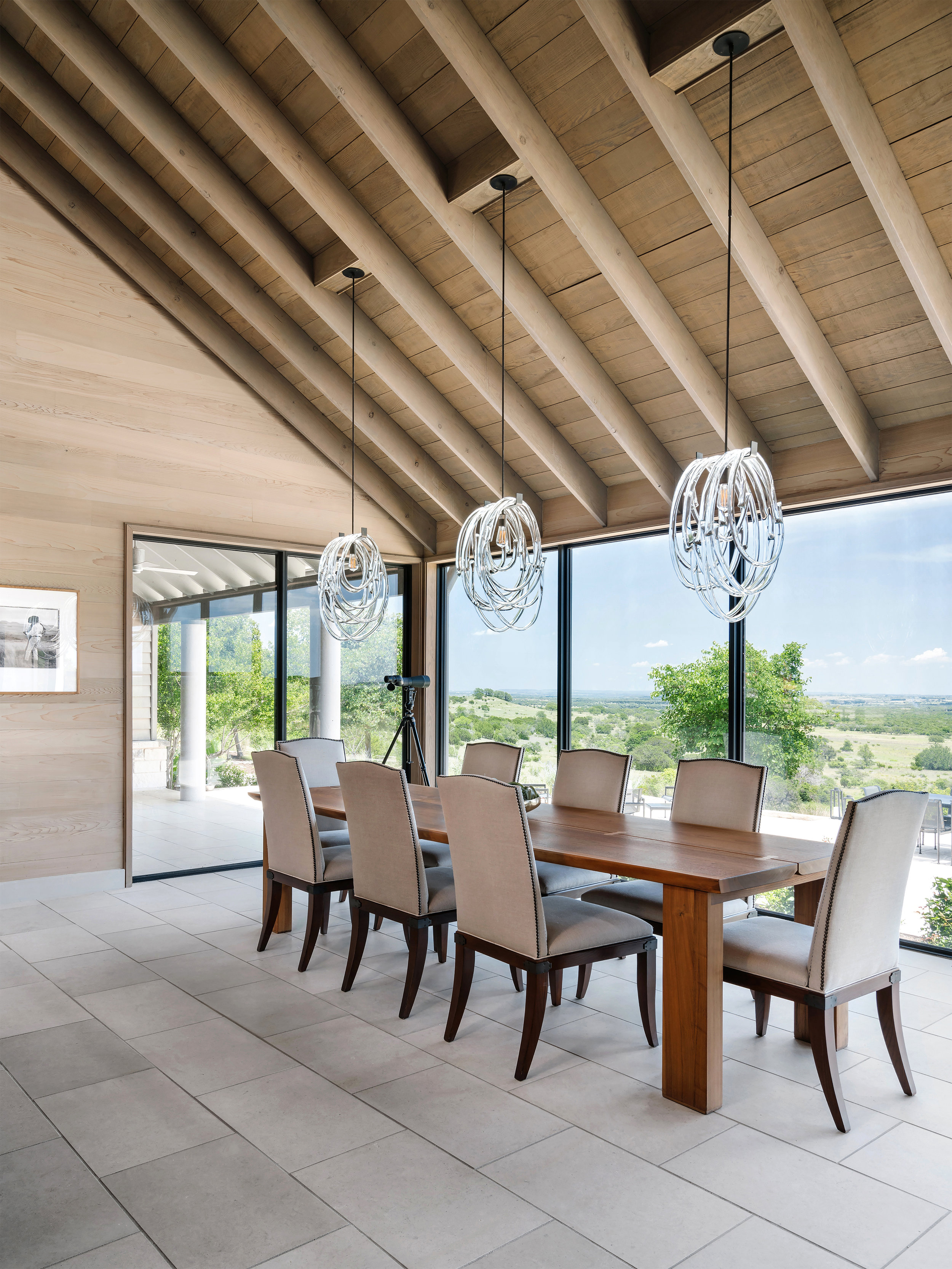 Dining room in a Lampassas ranch home with a wooden table, eight chairs, contemporary pendant lights, and large windows with scenic views.
