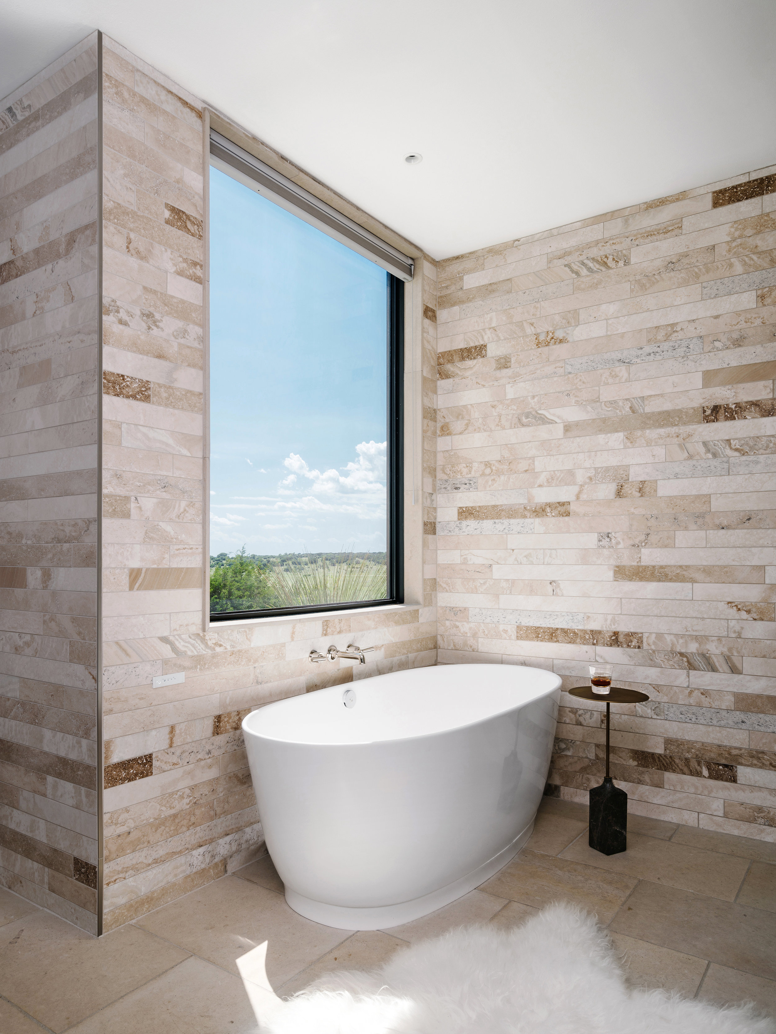 Bathroom in a Lampassas ranch home with a large bathtub, stone wall tiles, a window with scenic views, and a small table with a glass.