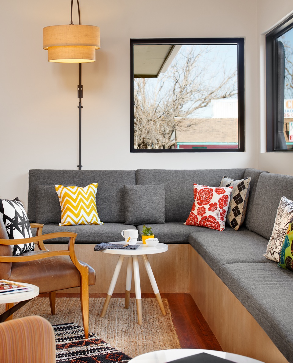 Cozy reception area in the Heywood Hotel with a gray sectional sofa, colorful pillows, wooden chair, small white table, and large windows.
