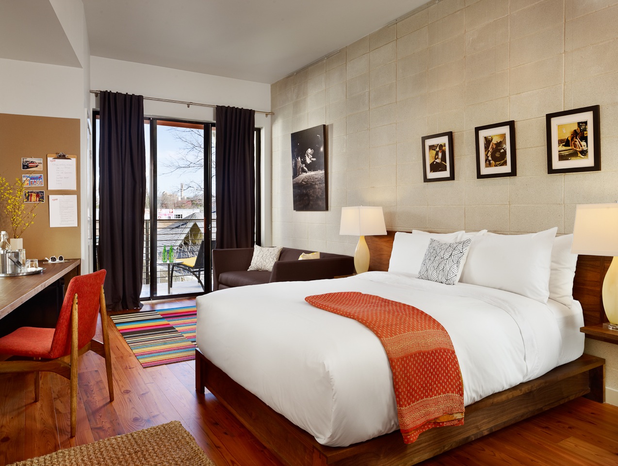 Cozy guest room in the Heywood Hotel with a king-size bed, colorful striped rug, wooden floors, desk, sofa, balcony, and framed artwork on the wall.