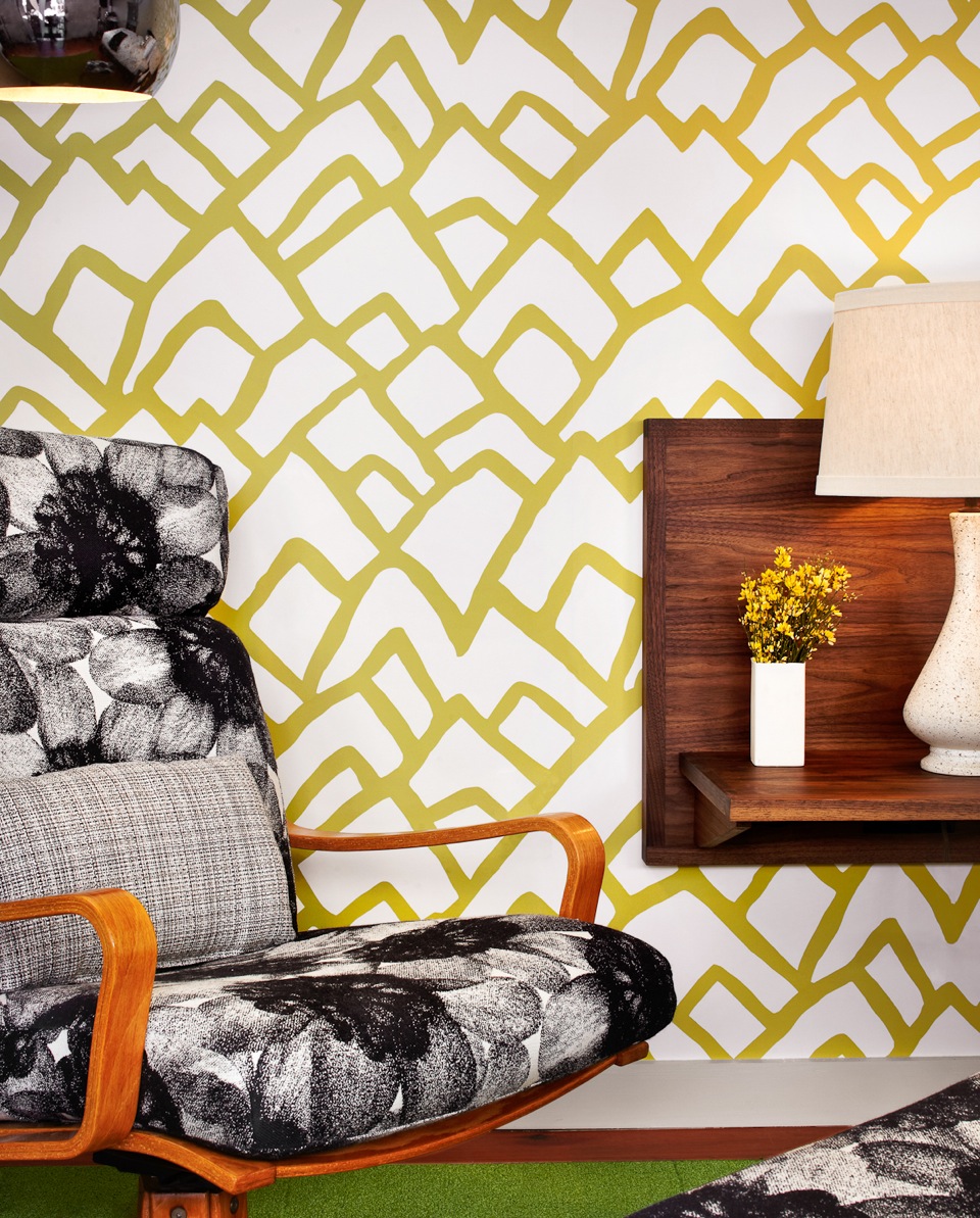 Mid-century modern chair with black-and-white floral pattern, geometric wallpaper, and wooden shelf with lamp and vase.