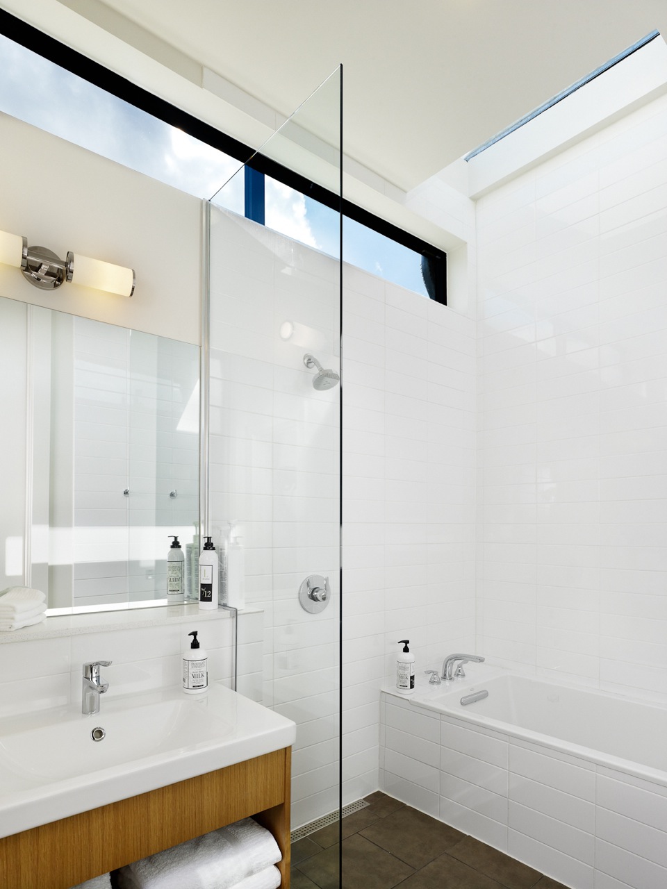 Bathroom in the Heywood Hotel with white tiles, a bathtub, a shower with a glass divider, a sink with wooden cabinet, and a mirror.