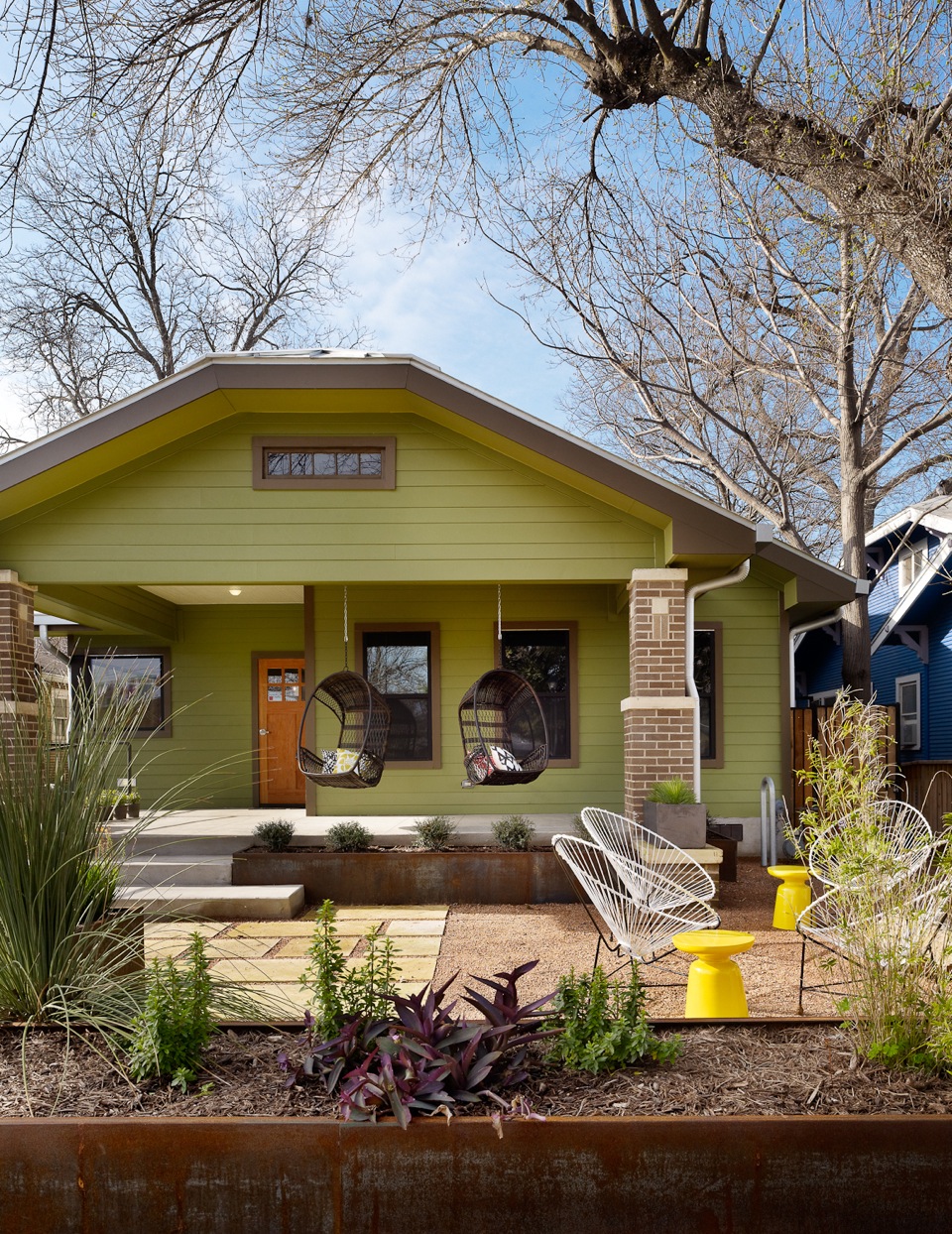 Exterior of the Heywood Hotel with a porch, two hanging chairs, and modern outdoor patio furniture, framed by leafless trees.