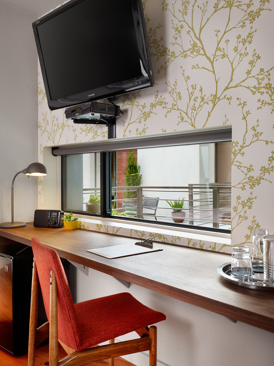A workspace in a guest room in the Heywood Hotel with a red chair, wooden desk, TV on the wall, desk lamp, and window with potted plants outside.