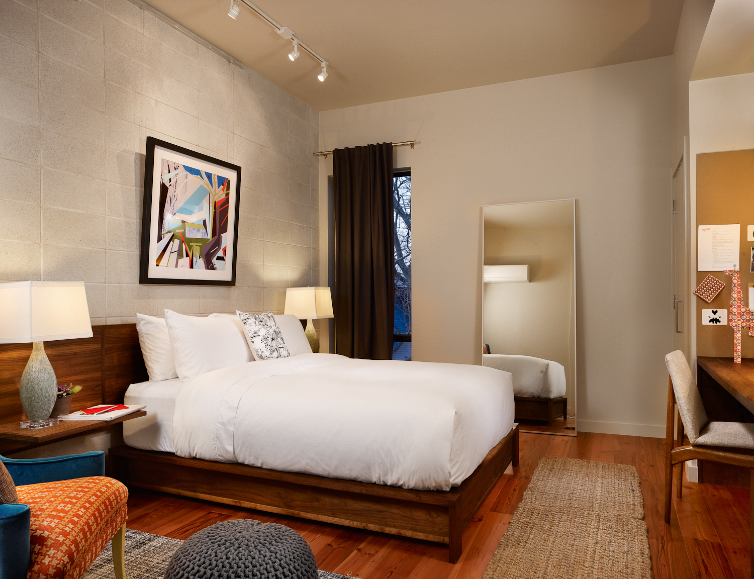 Guest room in the Heywood Hotel with a large bed, white bedding, colorful artwork on the wall, a large mirror, and a cozy seating area.