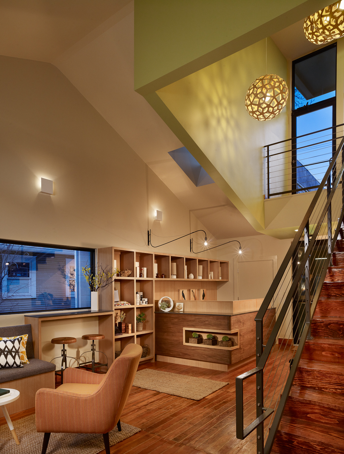 Reception area in the Heywood Hotel with wooden flooring, orange armchair, shelves, staircase, and decorative light fixtures hanging from ceiling.