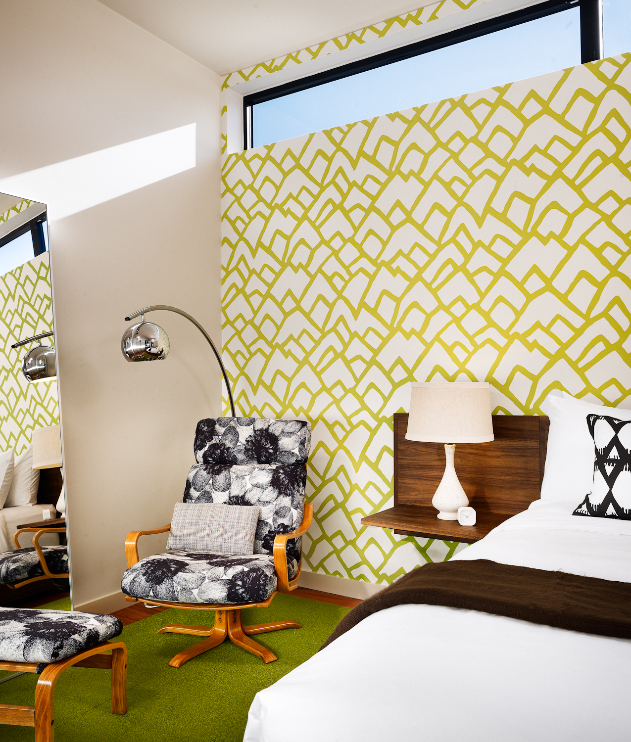 Guest room in the Heywood Hotel with a patterned accent wall, a chair and lamp beside a bed, and a green carpet.