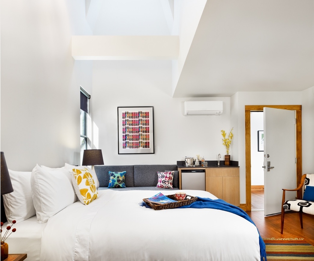 A bright guest room in the Heywood Hotel with a large bed, colorful pillows, a kitchenette, and a Mid-century modern chair.