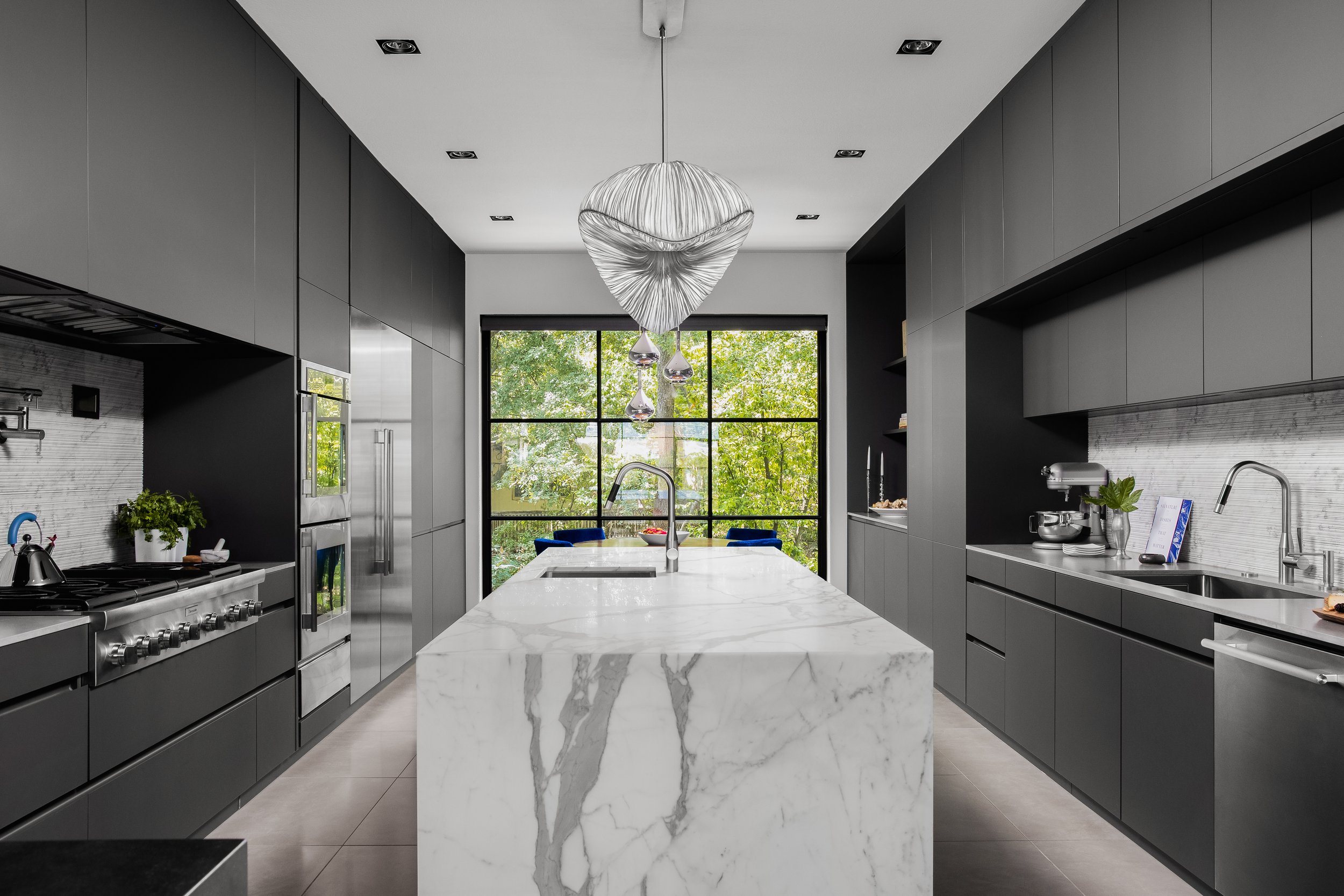 Kitchen in the Mediterranean Modern with dark cabinetry, white marble island, stainless steel appliances, and large central window.