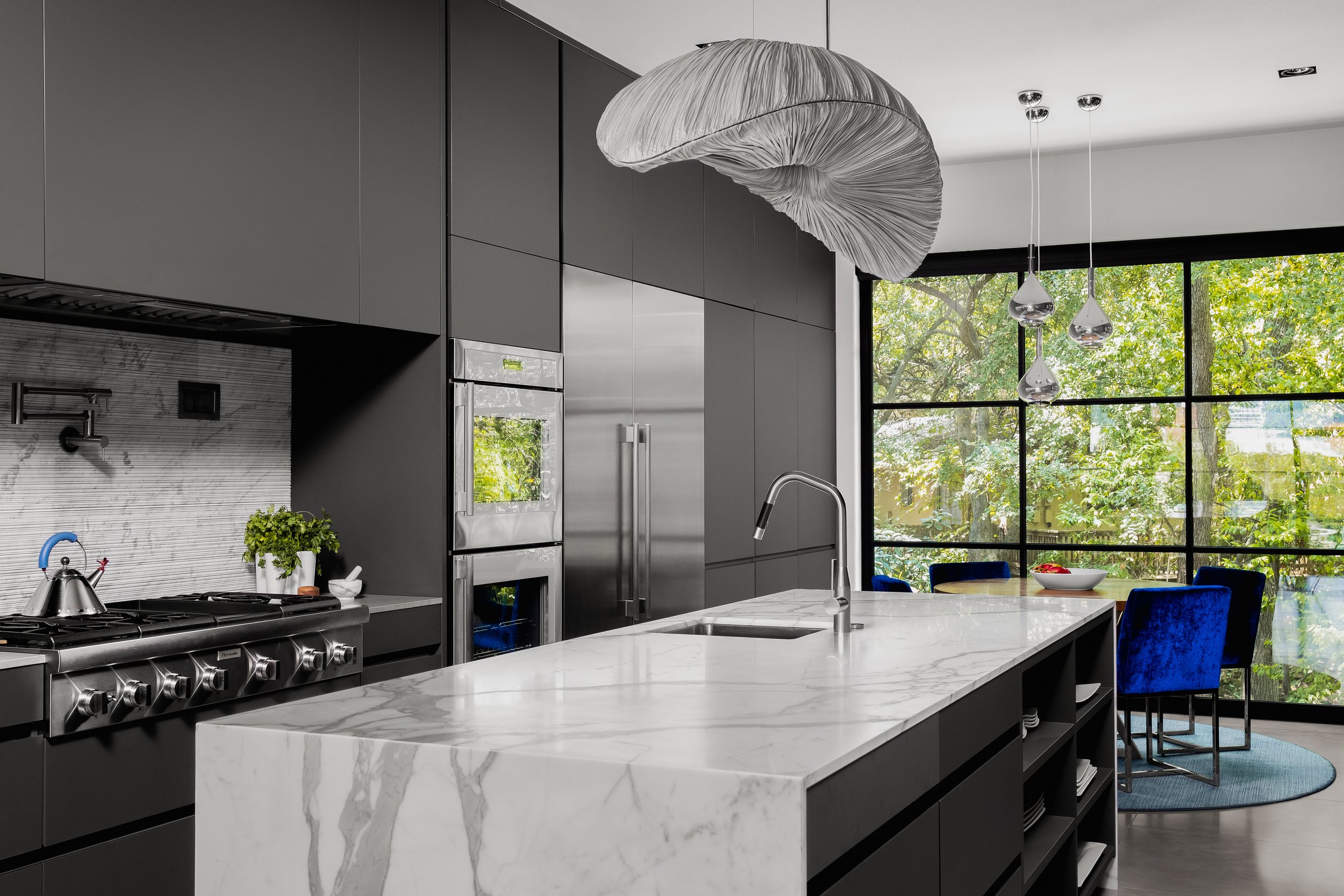 Kitchen in the Mediterranean Modern with dark cabinetry, marble island, large pendant light, stainless steel appliances, and large windows.