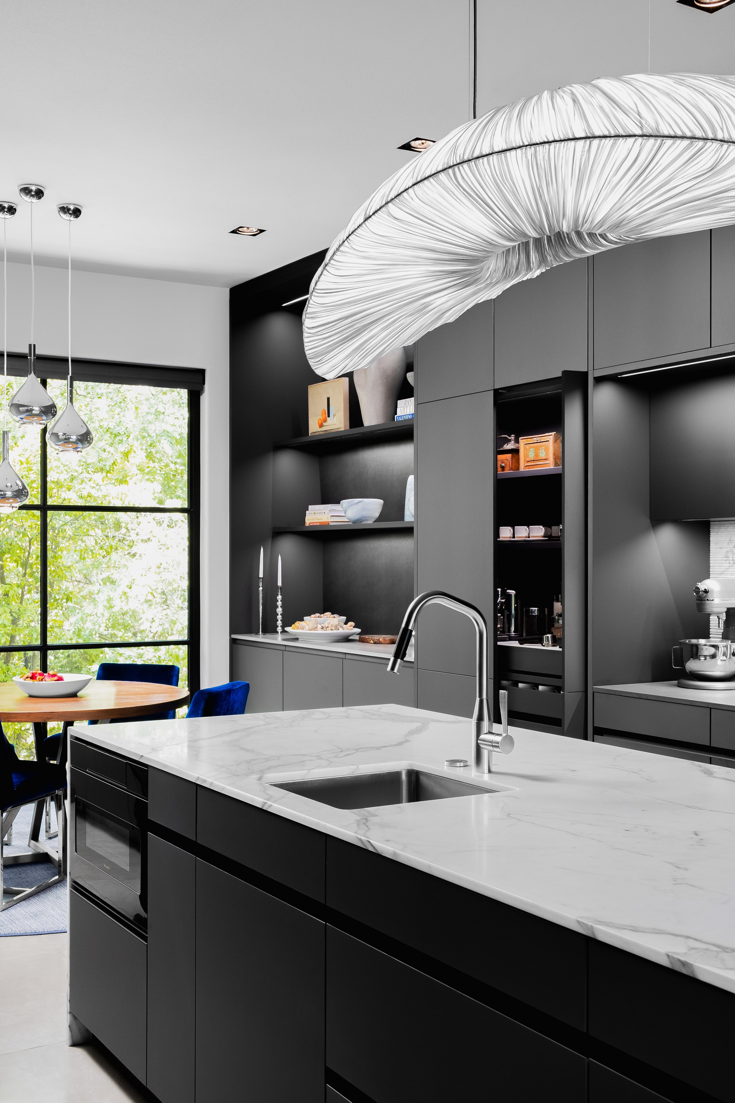 Kitchen in the Mediterranean Modern with dark cabinetry, a large marble island, stainless steel faucet, and a unique overhead light fixture.