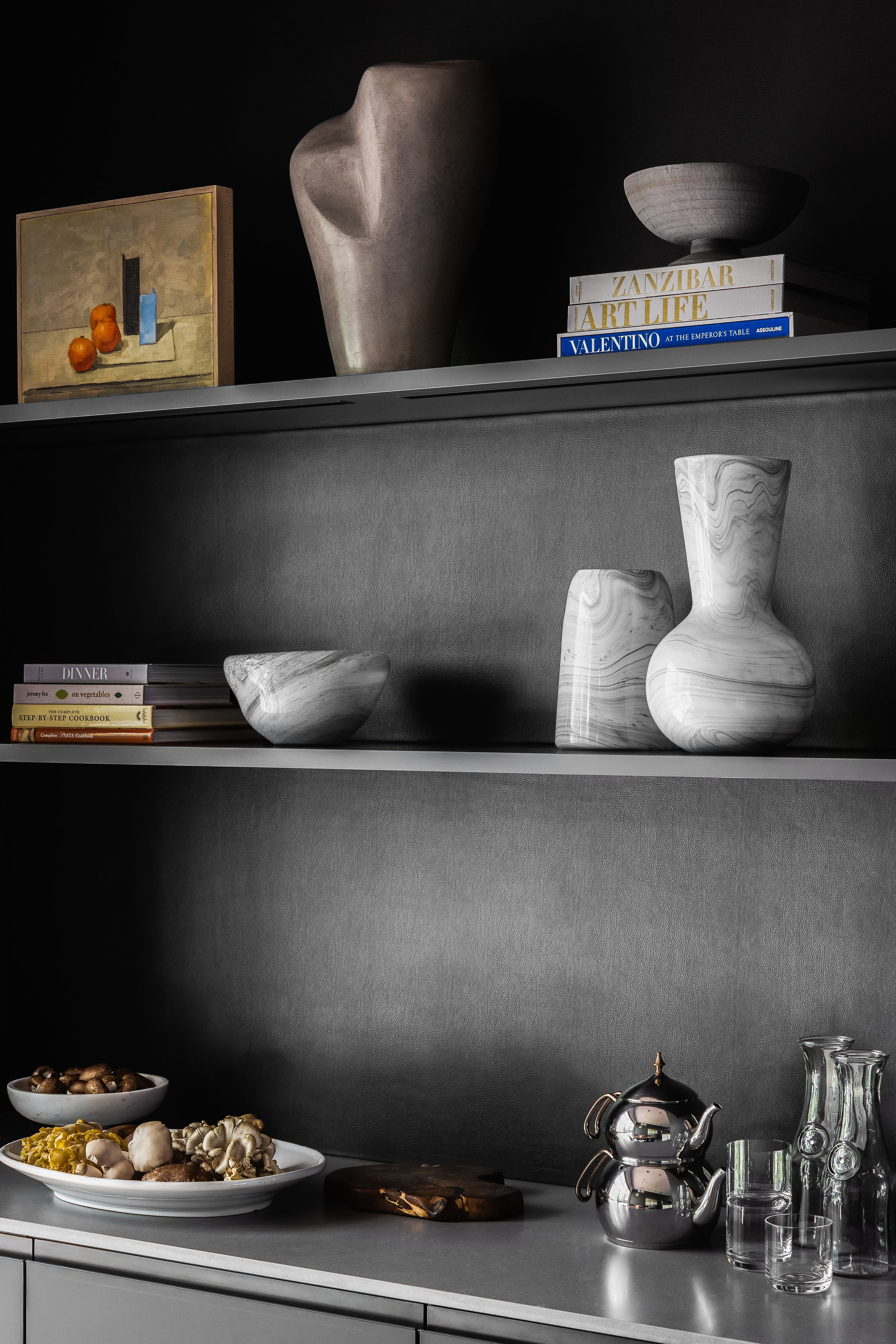 Kitchen shelf in the Mediterranean Modern with abstract sculptures, a tea set, fruits, and cookbooks neatly arranged on two dark shelves.