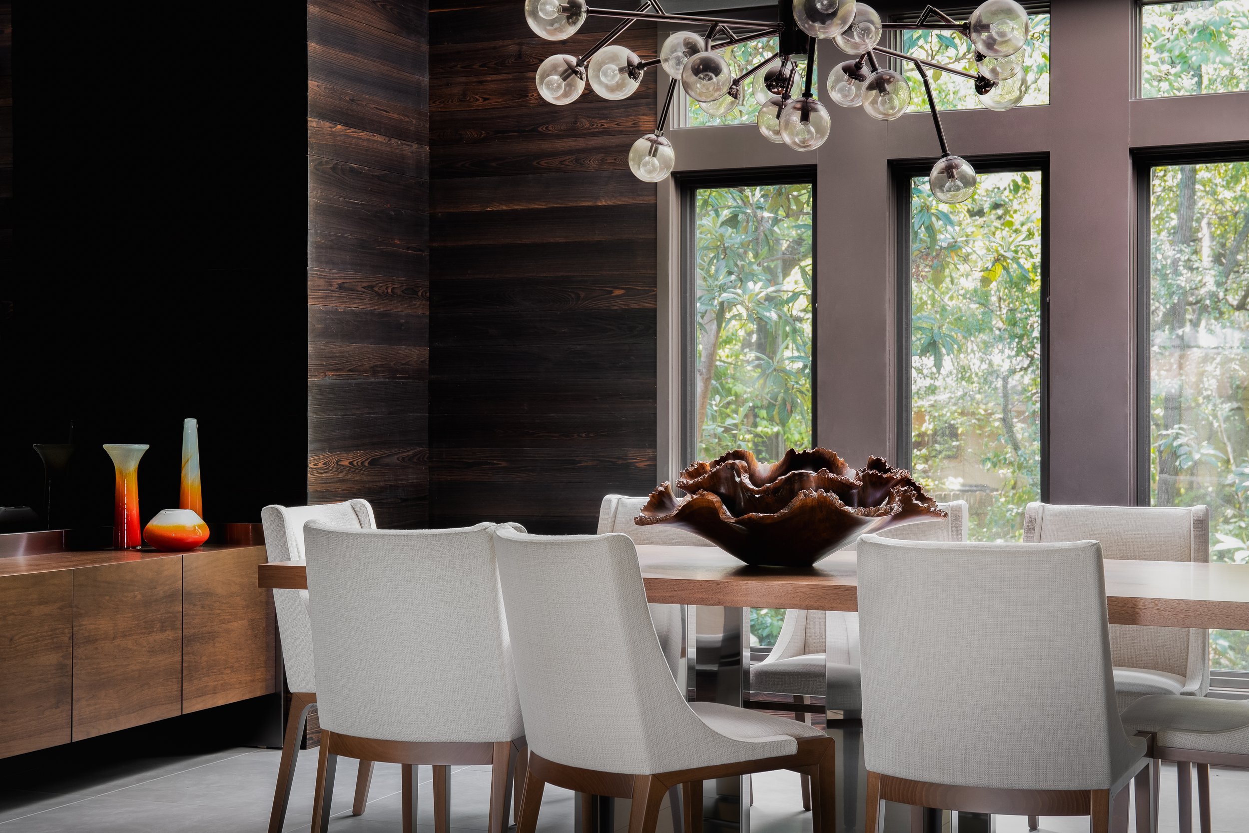 Dining room in the Mediterranean Modern with large windows, white chairs, a wooden table, a centerpiece sculpture, and a unique chandelier.