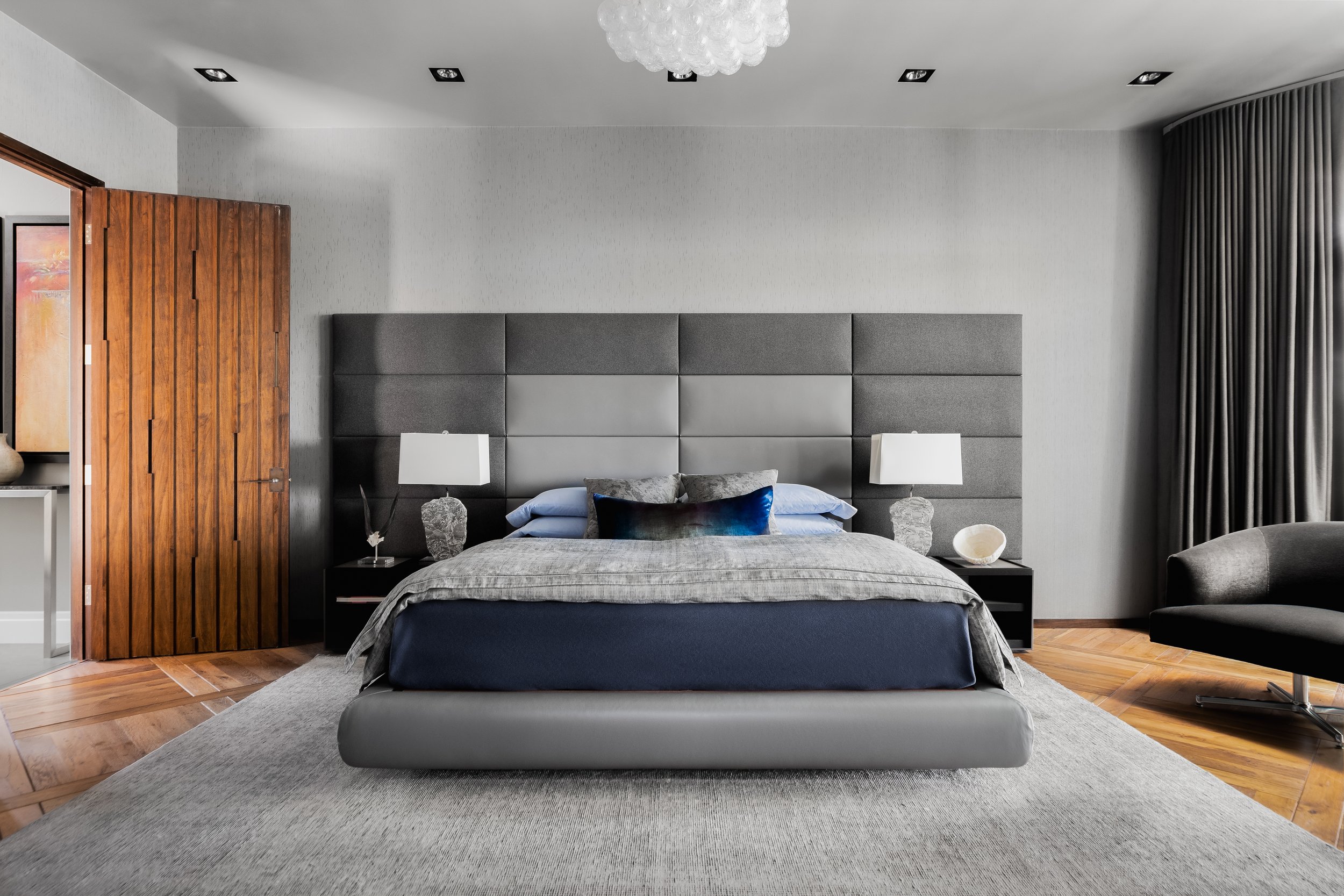 Bedroom in the Mediterranean Modern with wood accents, large upholstered headboard, and neutral tones. Bedside lamps and chair complete the decor.