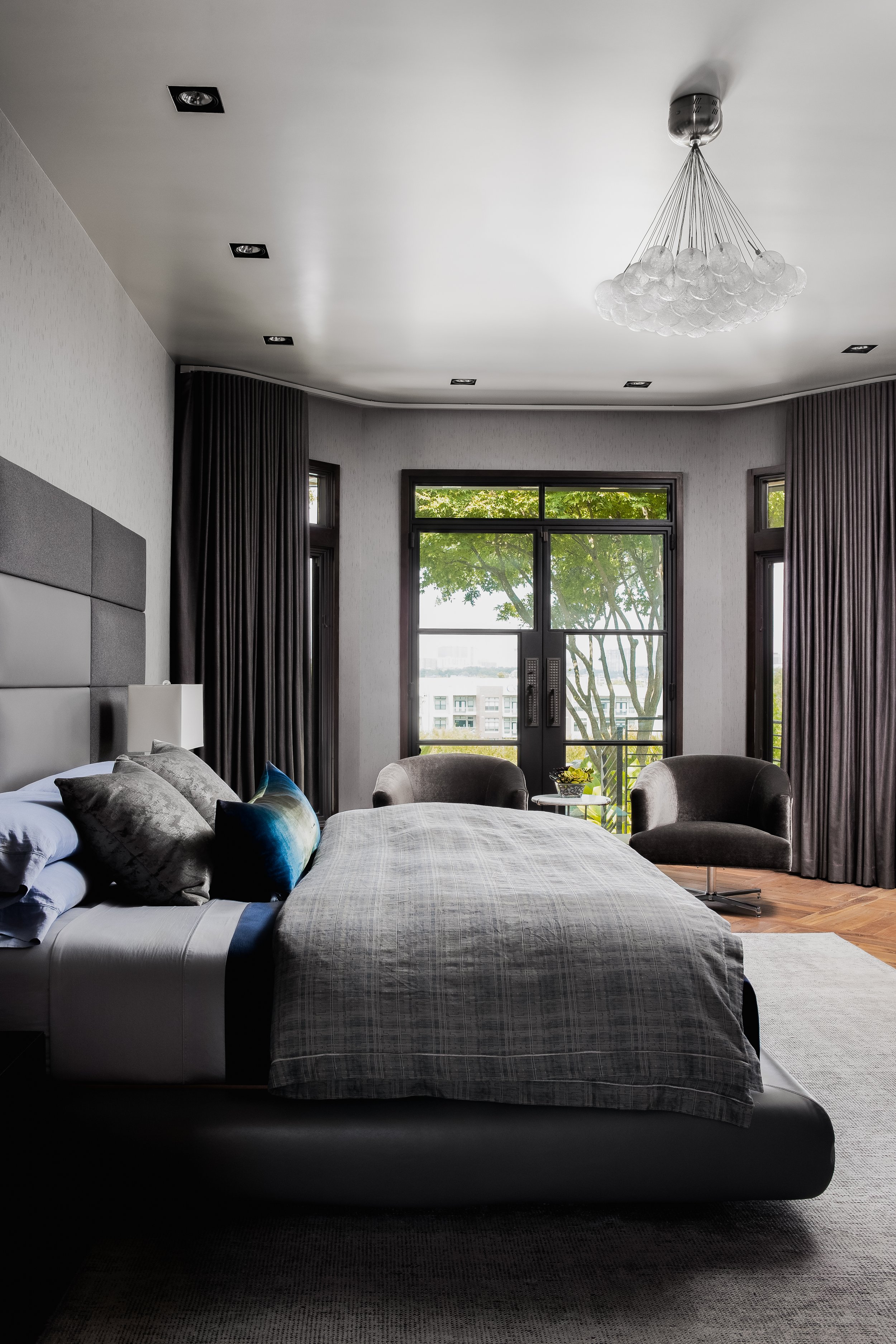 Bedroom in the Mediterranean Modern with a large bed, gray bedding, two armchairs, and floor-to-ceiling windows with dark curtains.