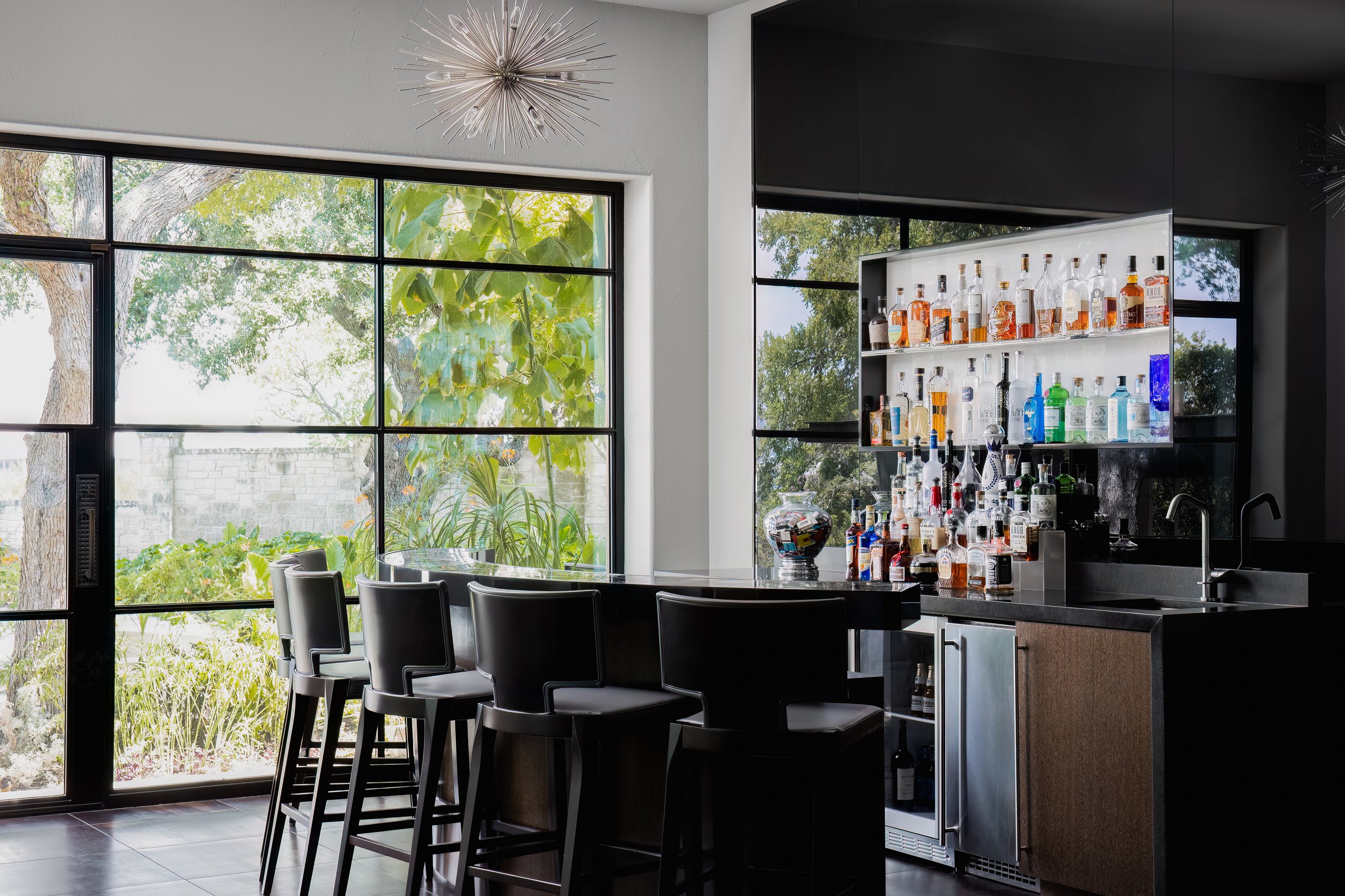 Home bar in the Mediterranean Modern with large windows, black chairs, sleek counter, and a well-stocked liquor shelf.