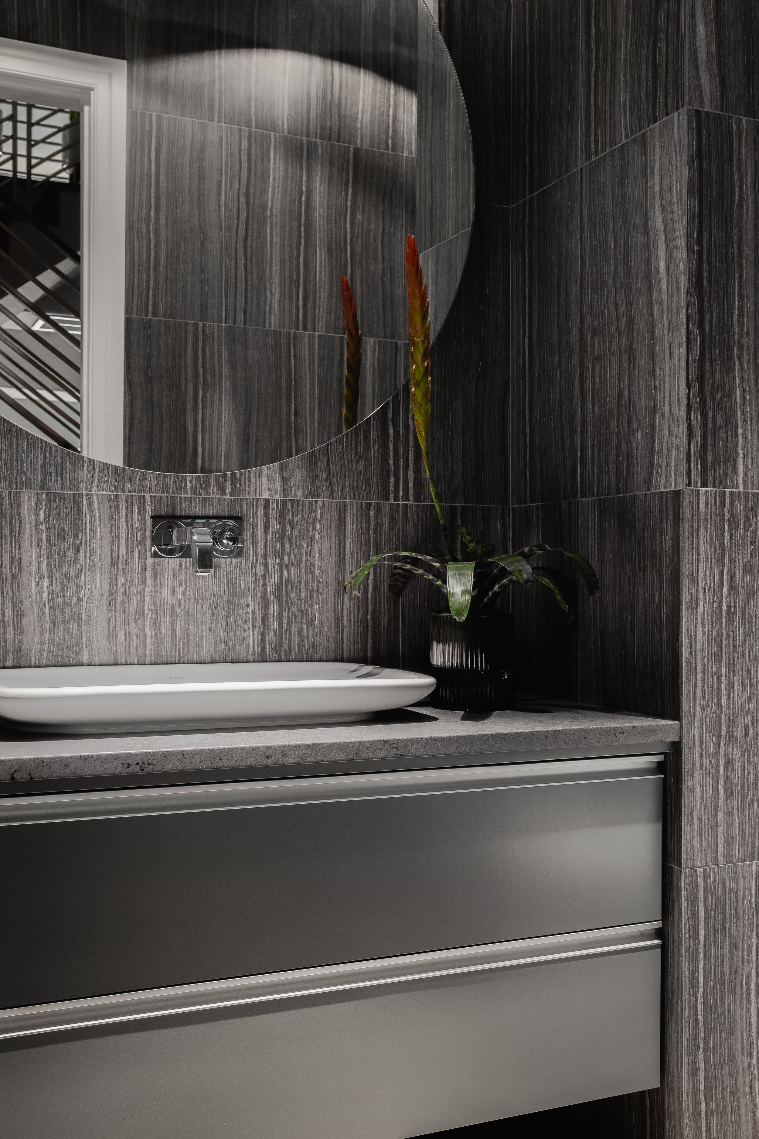 Bathroom in the Mediterranean Modern with dark grey marble walls, round mirror, white sink, and a decorative black vase.