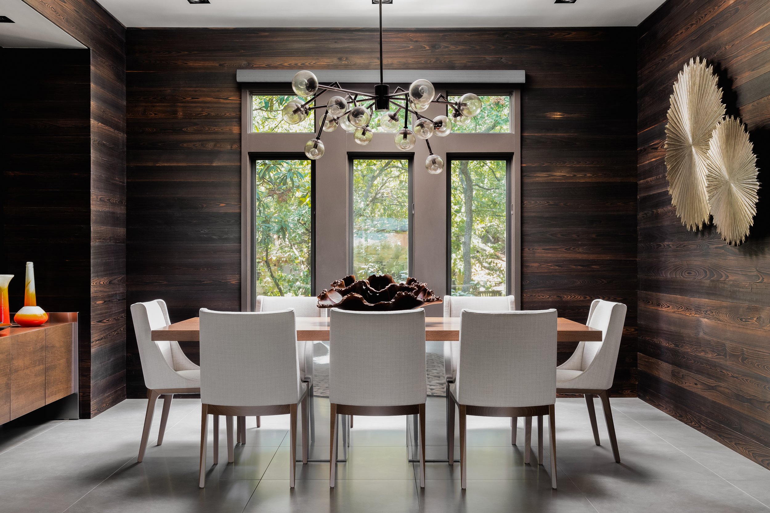 Dining room in the Mediterranean Modern with dark wood walls, a wooden table, white chairs, a spherical chandelier, and large windows.