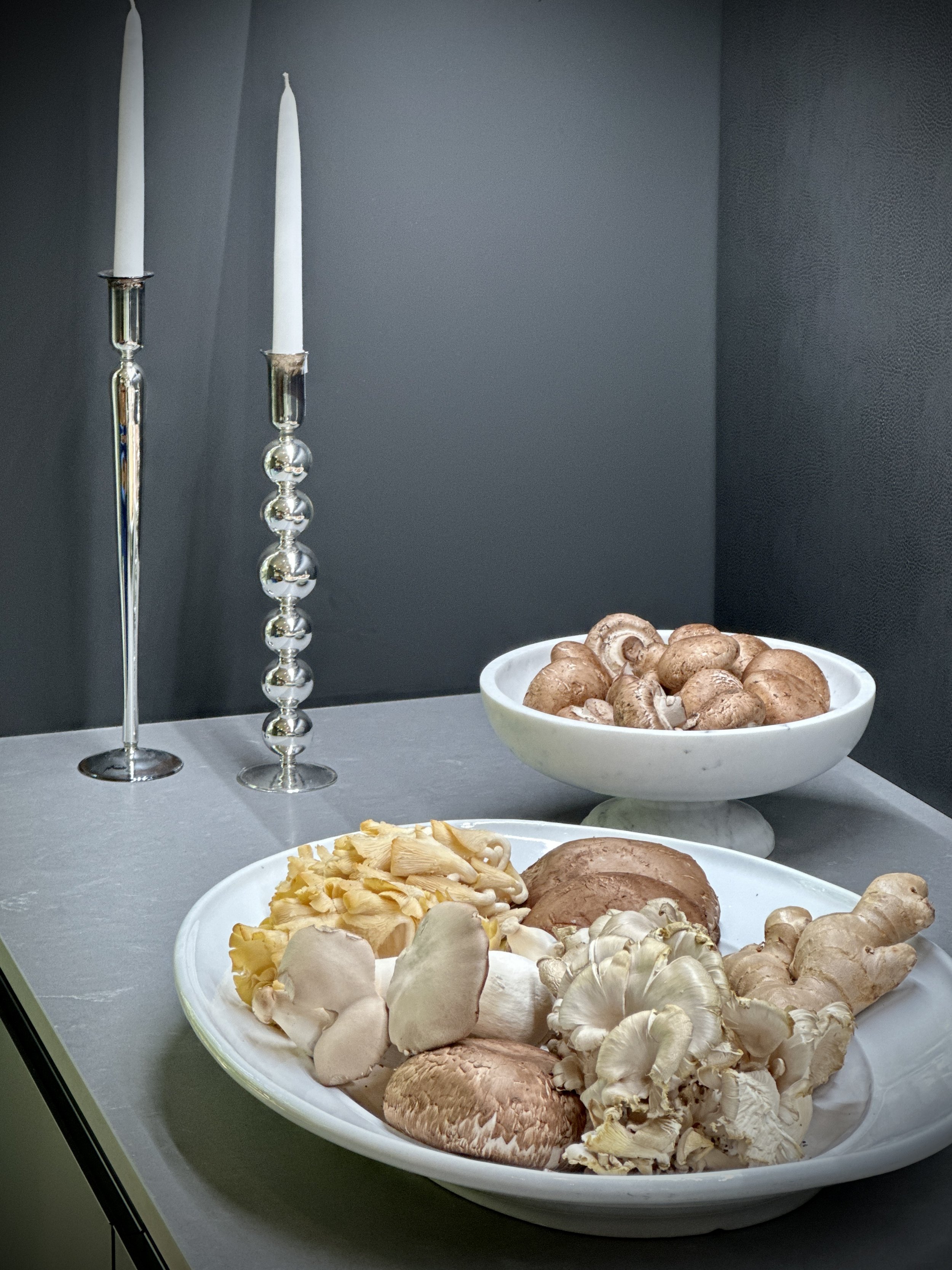 Plates of assorted mushrooms on a grey countertop, with tall, decorative candle holders in the background.
