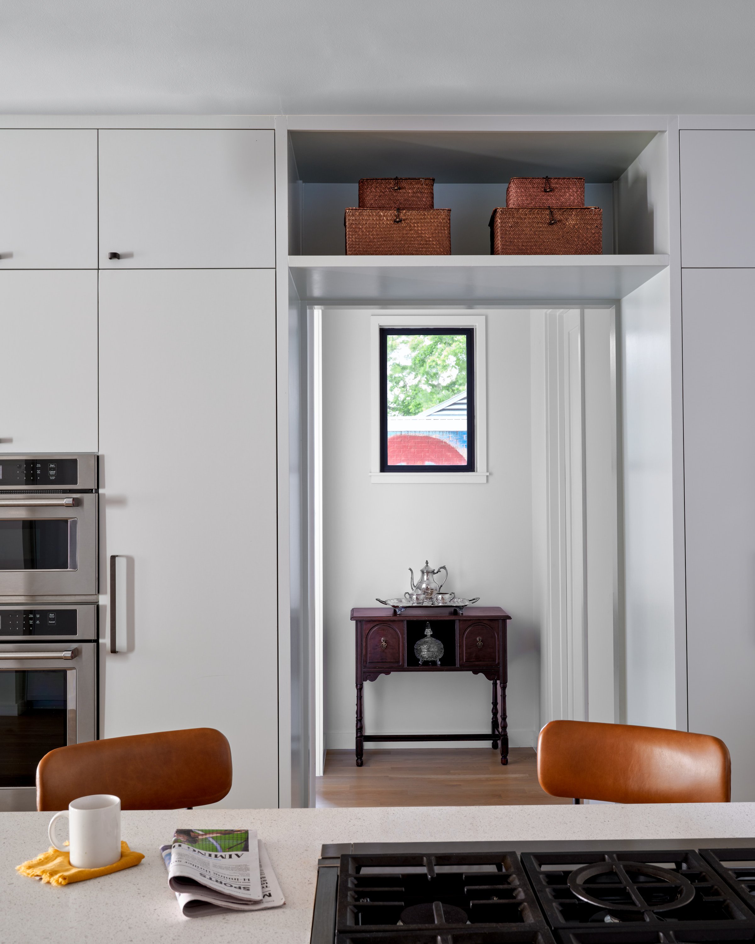 Kitchen in an East Side bungalow with a white countertop, gas stove, and wall ovens with a passageway leading to a window with a wooden table underneath.