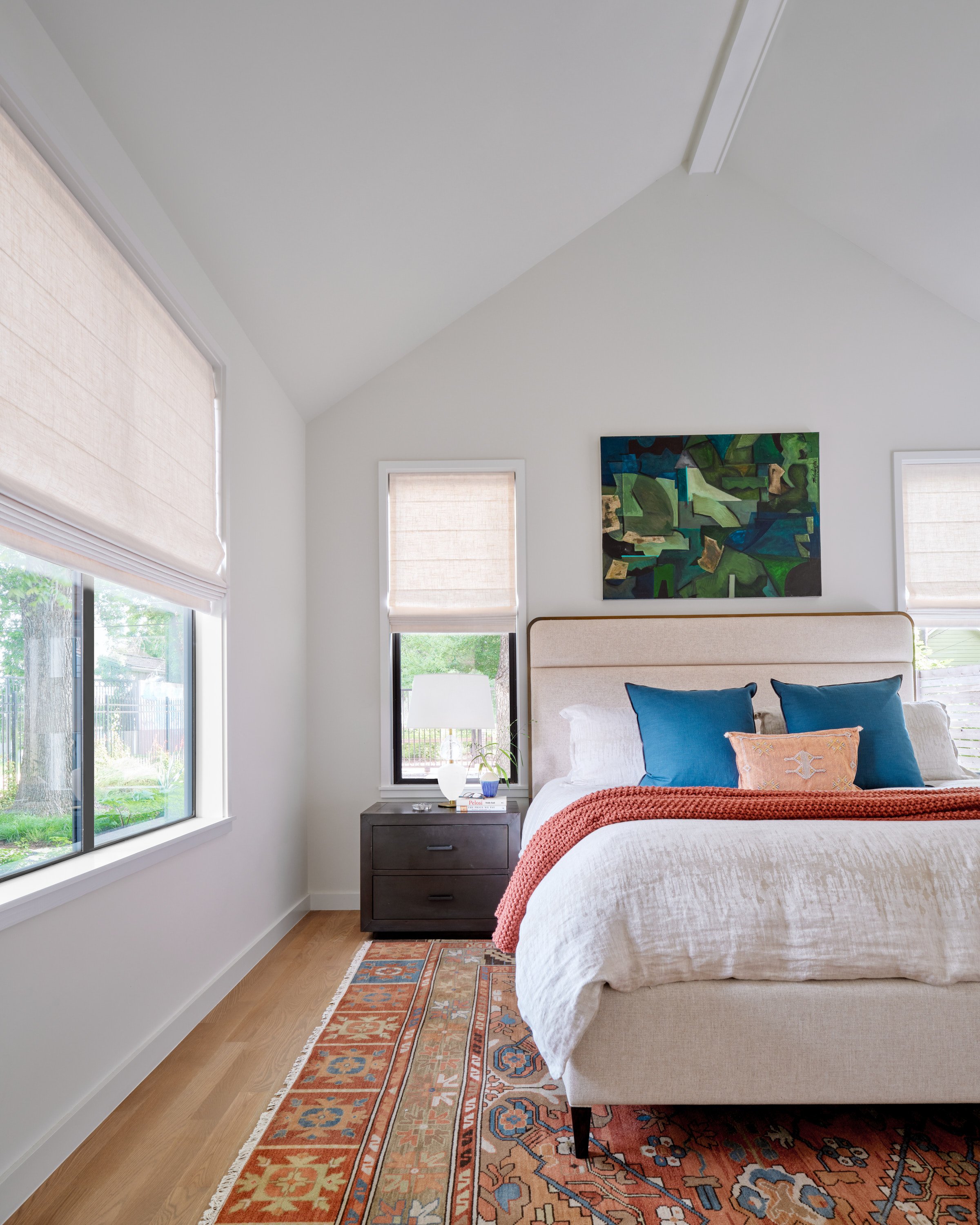 A bedroom in an East Side bungalow with a bed, blue and orange pillows, three windows, a nightstand, and abstract artwork above the headboard.