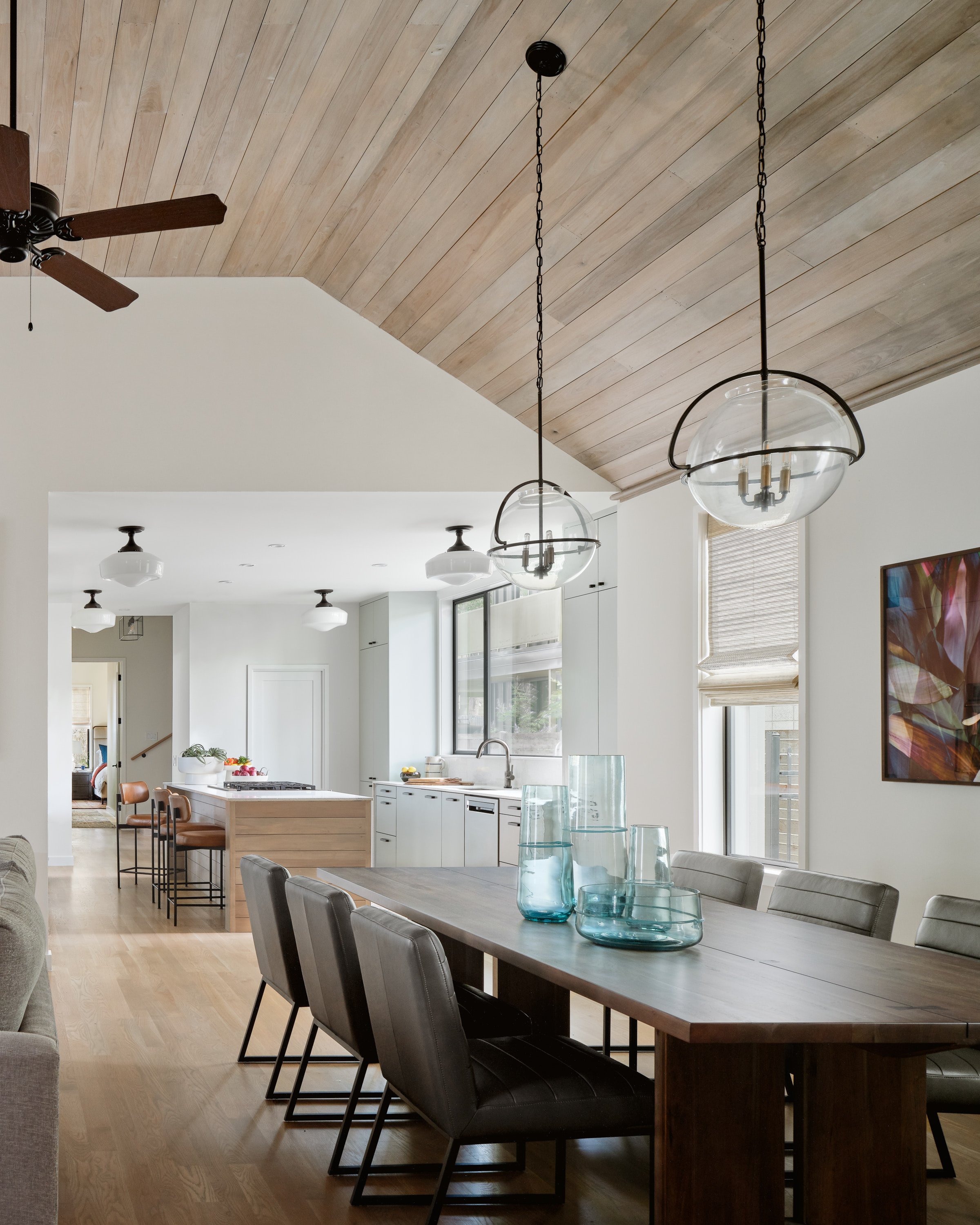 Dining area in an East Side bungalow with a wooden ceiling, pendant lights, a large table with gray chairs, and a spacious kitchen in the background.