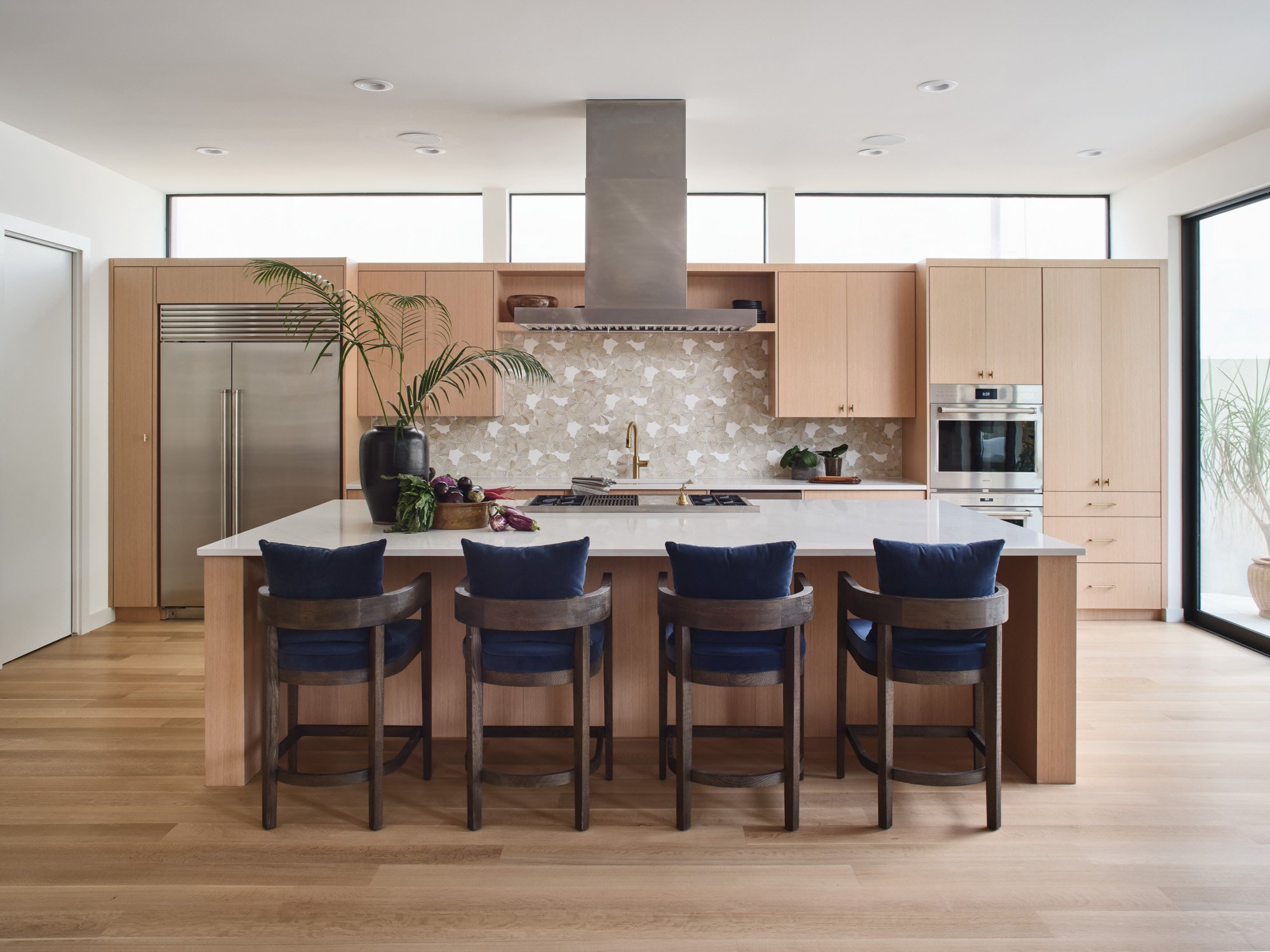 Kitchen in a Barton Creek home with an island, blue cushion bar stools, stainless steel appliances, and light wood cabinets and flooring.
