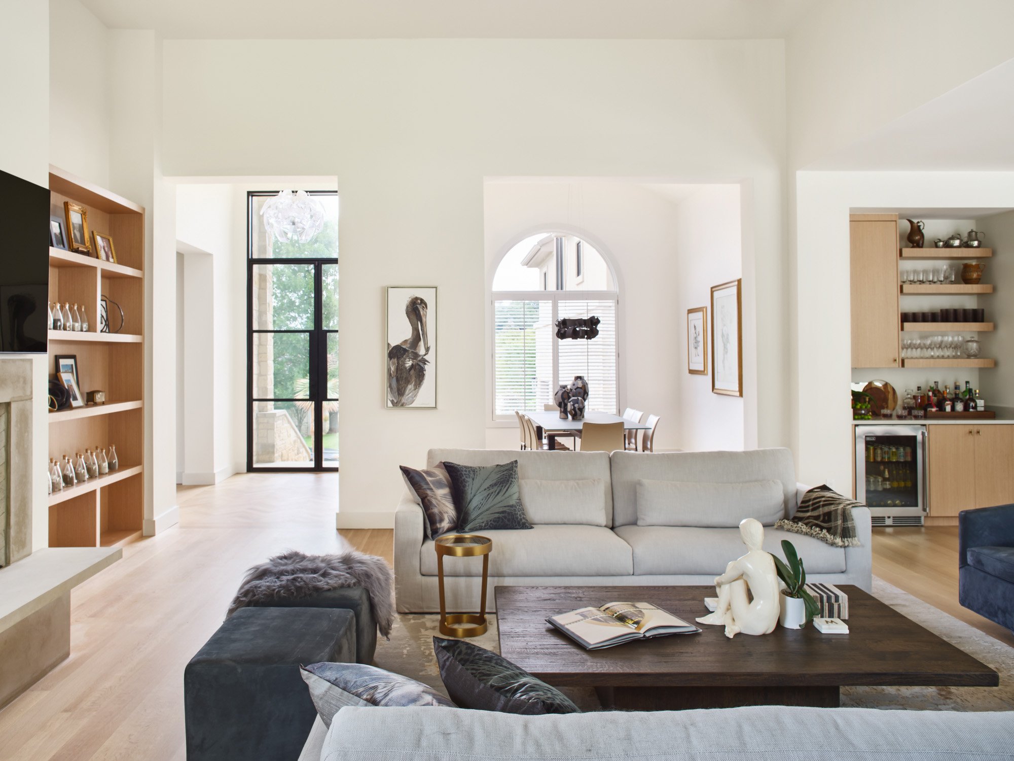 Living room in a Barton Creek home with neutral tones, a large sofa, wooden table, and open pathways to dining and kitchen areas, with lots of art on the walls and shelves.