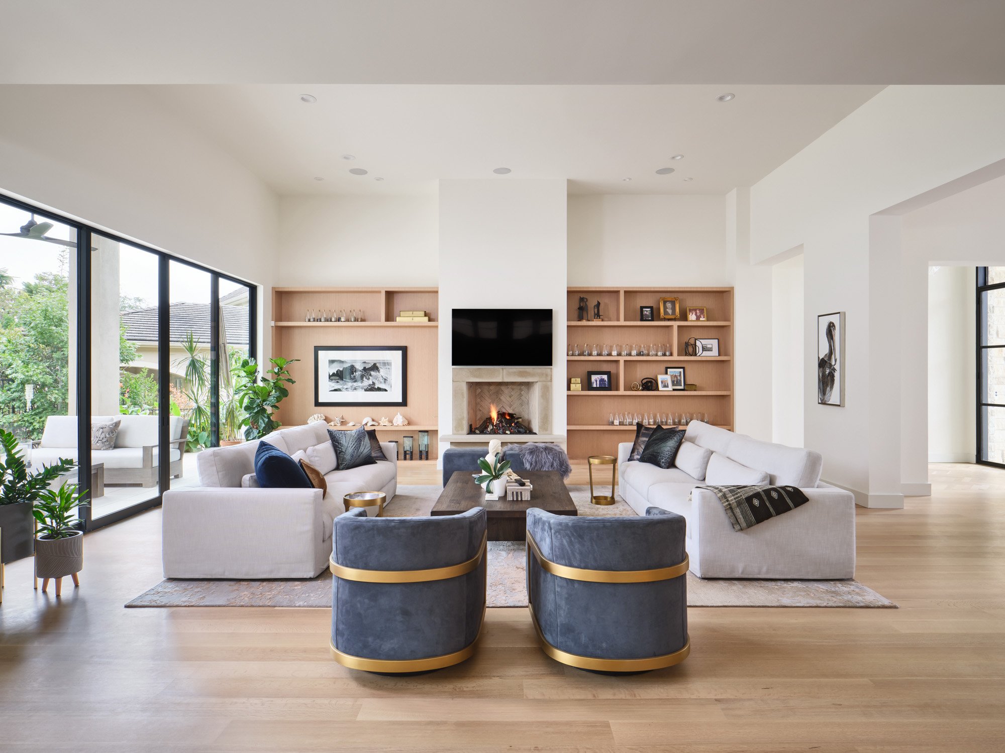Living room in a Barton Creek home with white sofas, gray armchairs, wooden shelves, a fireplace, and large windows opening to a patio.