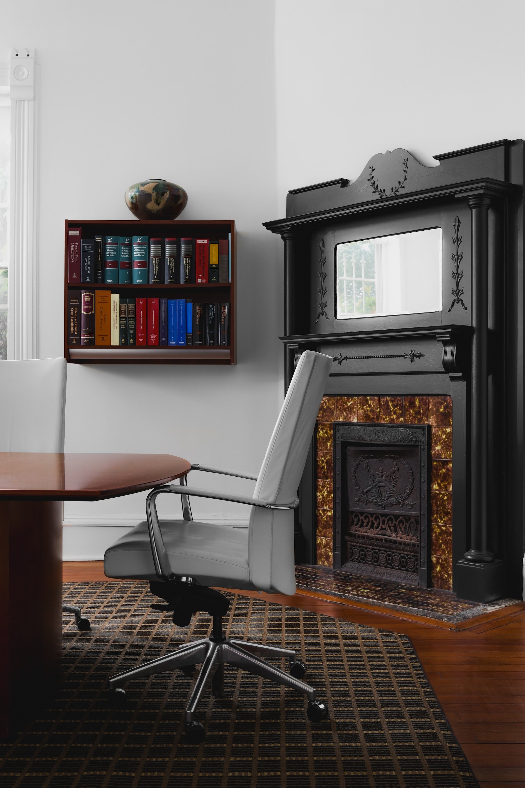 Office space in a law firm with a modern chair, wooden desk, black fireplace, mirror, bookshelf with books, and a window.
