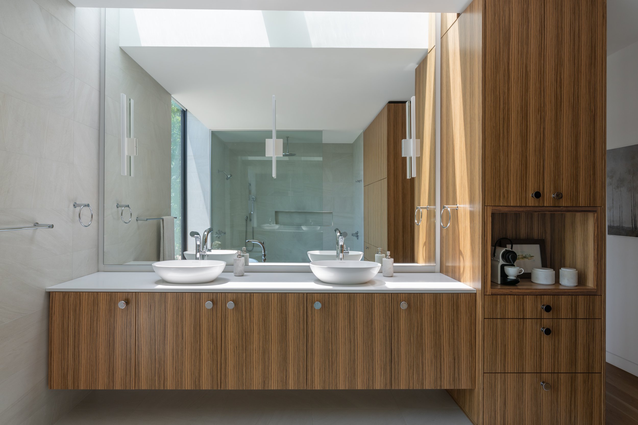 Bathroom in a Laurel Canyon home with wood cabinetry, double sinks, large mirror, modern light fixtures, and a glass walk-in shower reflected in the background.