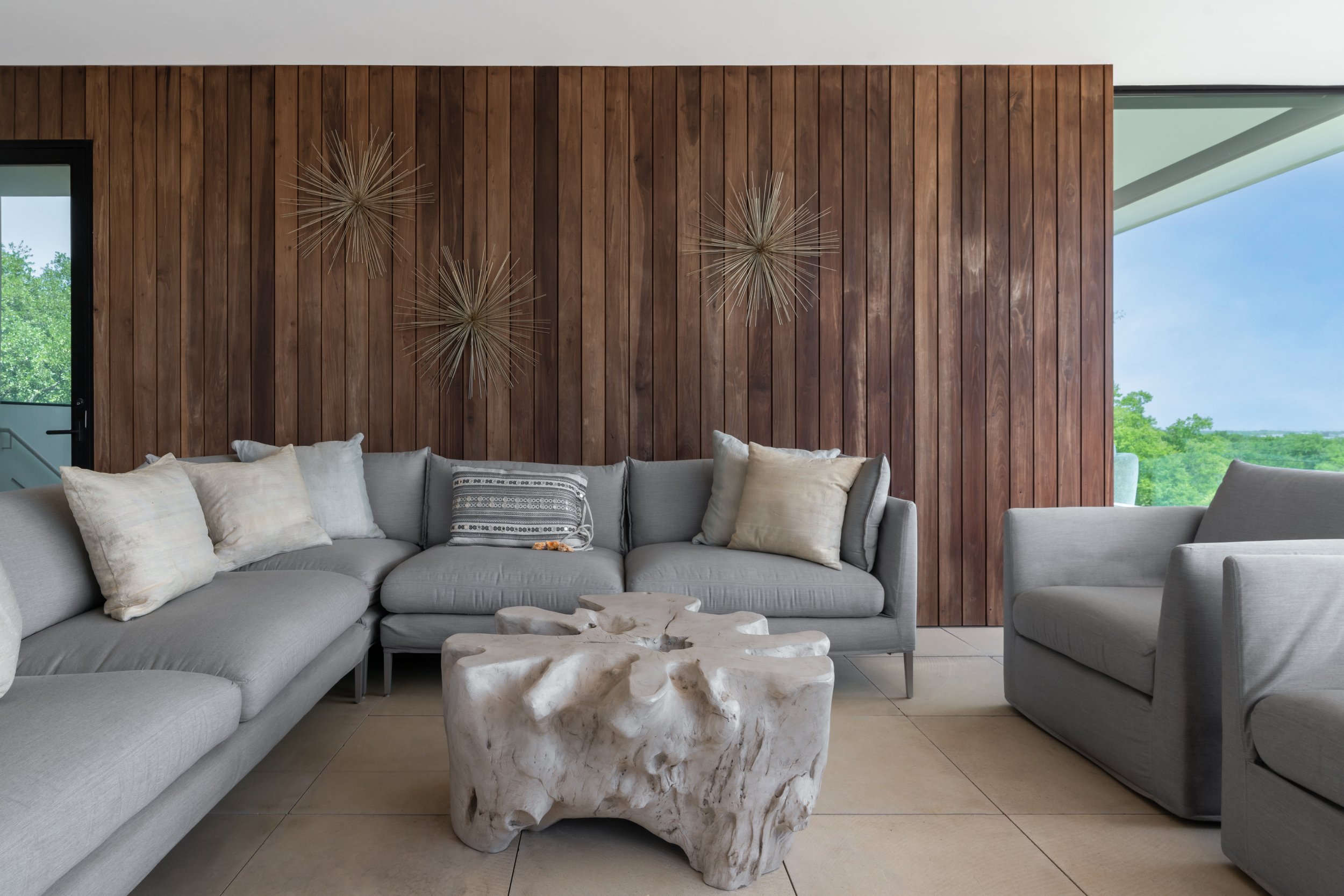 Living room in a Laurel Canyon home with grey sofas, a tree stump shaped coffee table, and wooden panel wall decor with large windows providing an outdoor view.